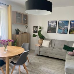 Olena Ryzhkova - Jasne wnętrze salonu z drewnianym stołem z bukietem fioletowych kwiatów, szarą sofą narożną, czarnym okrągłym żyrandolem i trzema obrazami górskimi na ścianie.