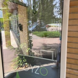 Olena Ryzhkova - Wysokie okno biurowe z widocznym banerem reklamowym firmy OYZO Dystrybucja, odbijające otoczenie zewnętrzne z drzewami i chodnikiem.
