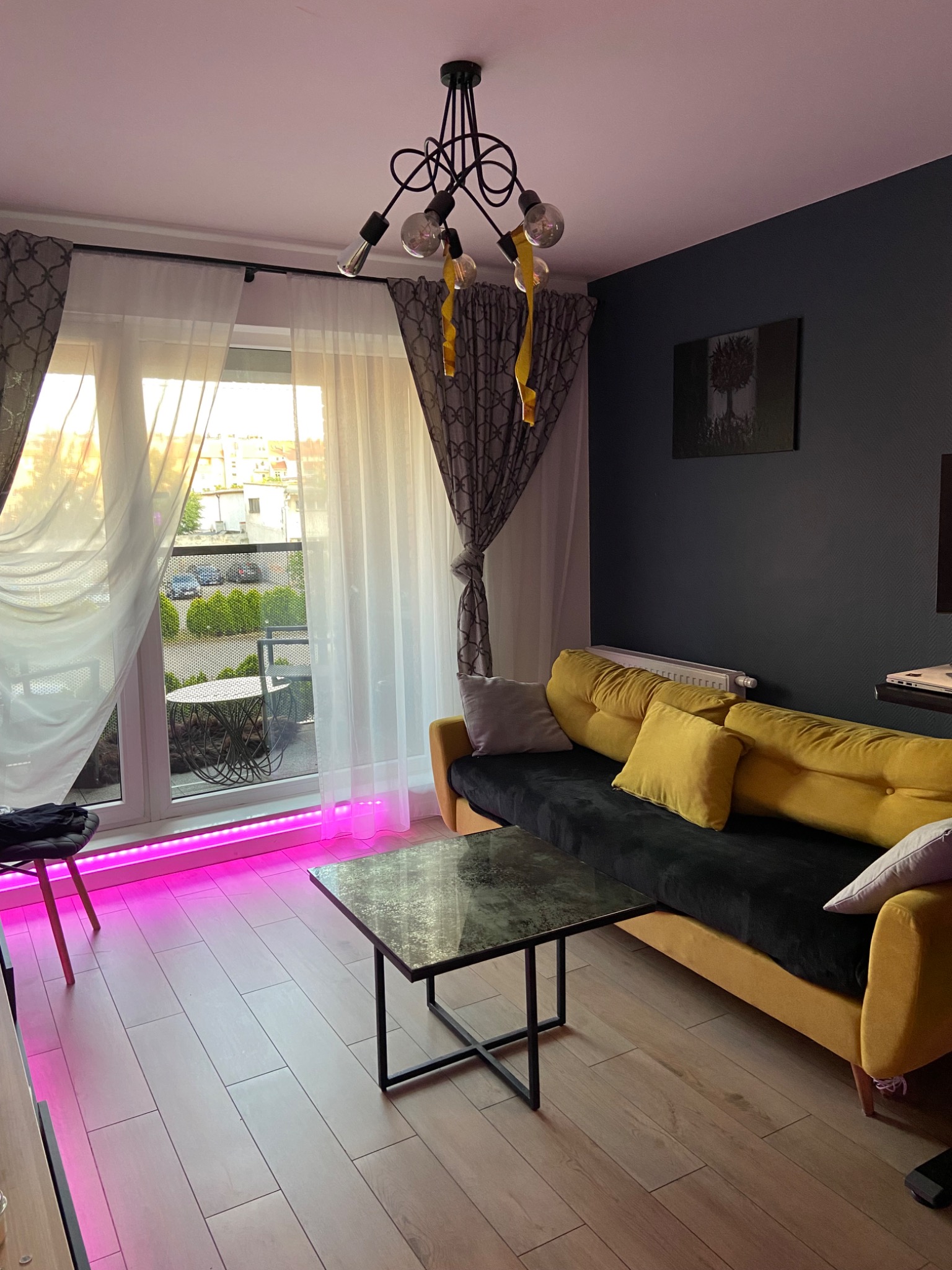 Nowoczesny salon z żółtą sofą, marmurkowym stolikiem kawowym i oknem balkonowym z widokiem na zieleń, oświetlony listwą LED w kolorze fuksji.
