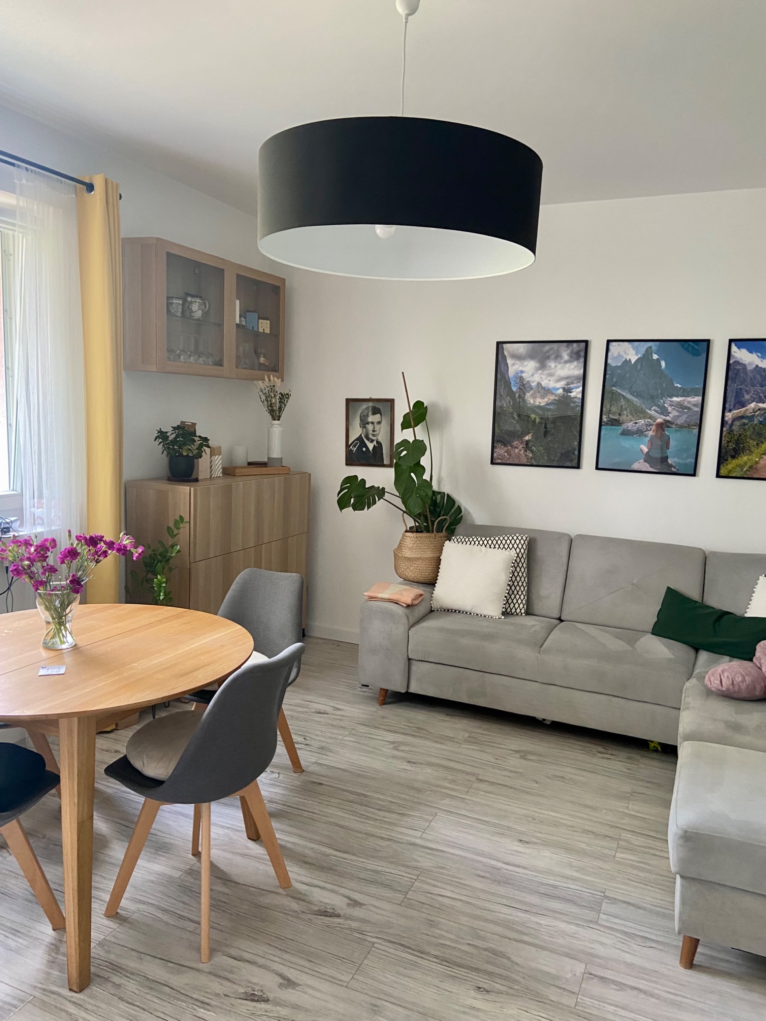 Jasne wnętrze salonu z drewnianym stołem z bukietem fioletowych kwiatów, szarą sofą narożną, czarnym okrągłym żyrandolem i trzema obrazami górskimi na ścianie.