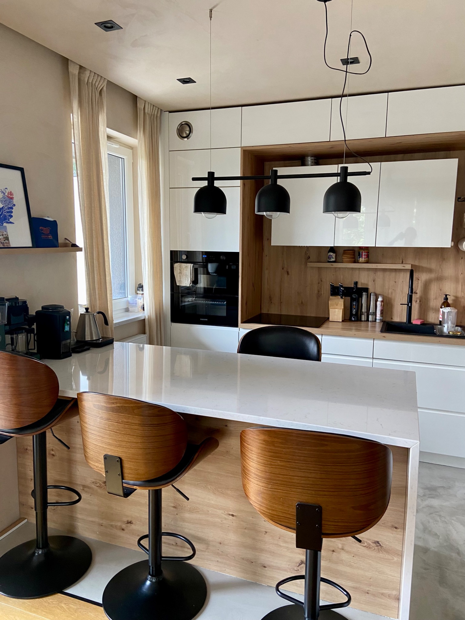 Nowoczesna kuchnia z wyspą i drewnianymi hokerami, minimalistyczne oświetlenie i zabudowa kuchenna w bieli i drewnie.