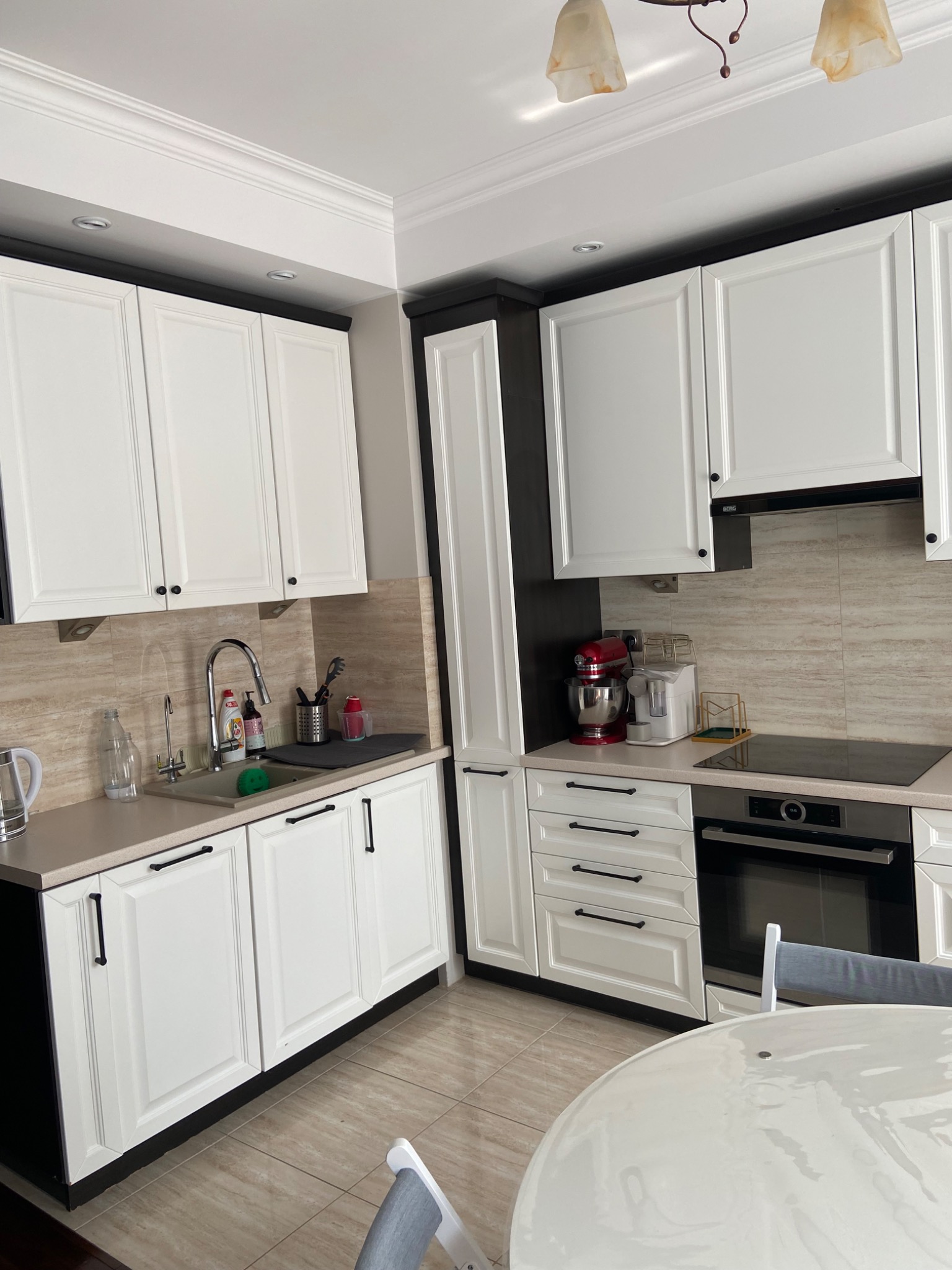 Jasna kuchnia z białymi szafkami i czarnymi akcentami, widok na zlew z płynem do naczyń, kuchenkę, piekarnik i mikser stojący na blacie.