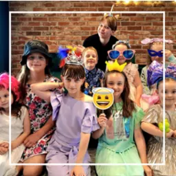 Grupa dzieci w różnorodnych przebraniach i nakryciach głowy, pozująca do zdjęcia na tle ceglanej ściany, z dziewczynką trzymającą rekwizyt emotikonu anioła.