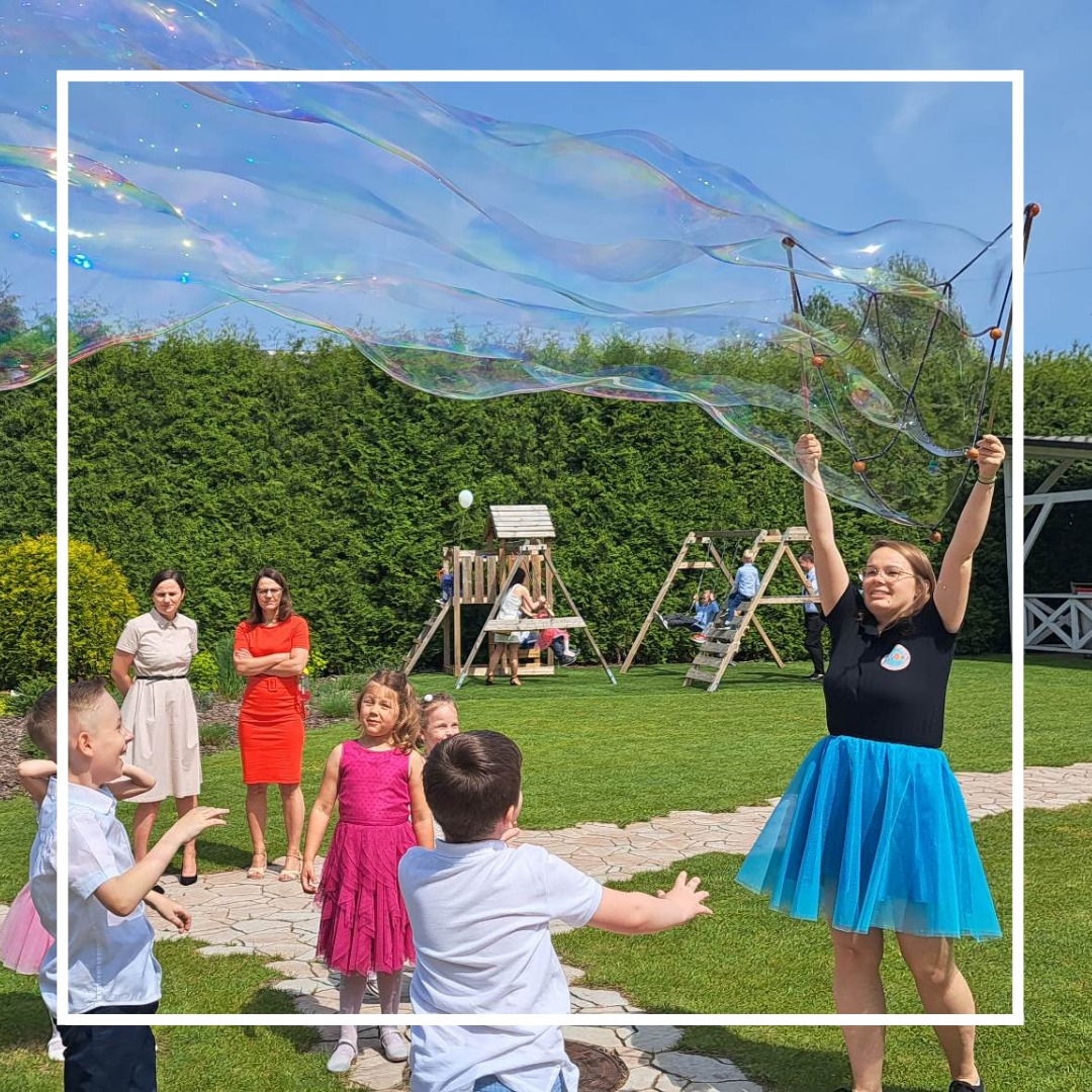 Animator w niebieskiej spódnicy tworzy gigantyczne bańki mydlane na trawniku podczas imprezy plenerowej dla dzieci, w tle huśtawki i dorośli.