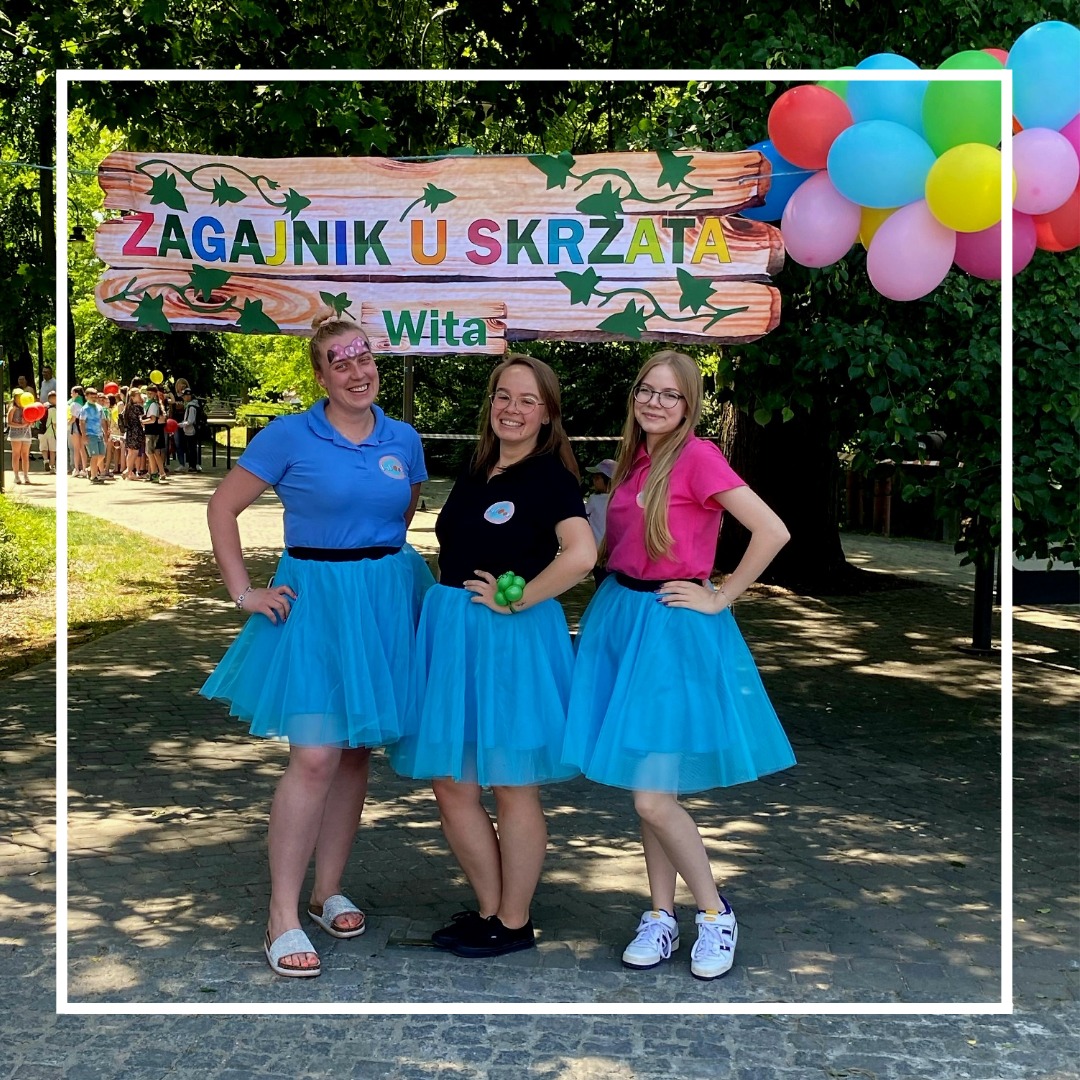 Trzy kobiety w kolorowych strojach (niebieskie i różowe koszulki oraz turkusowe spódniczki tutu) pozują przed drewnianym szyldem 'ZAGAJNIK U SKRZATA', z balonami w tle, prawdopodobnie podczas...