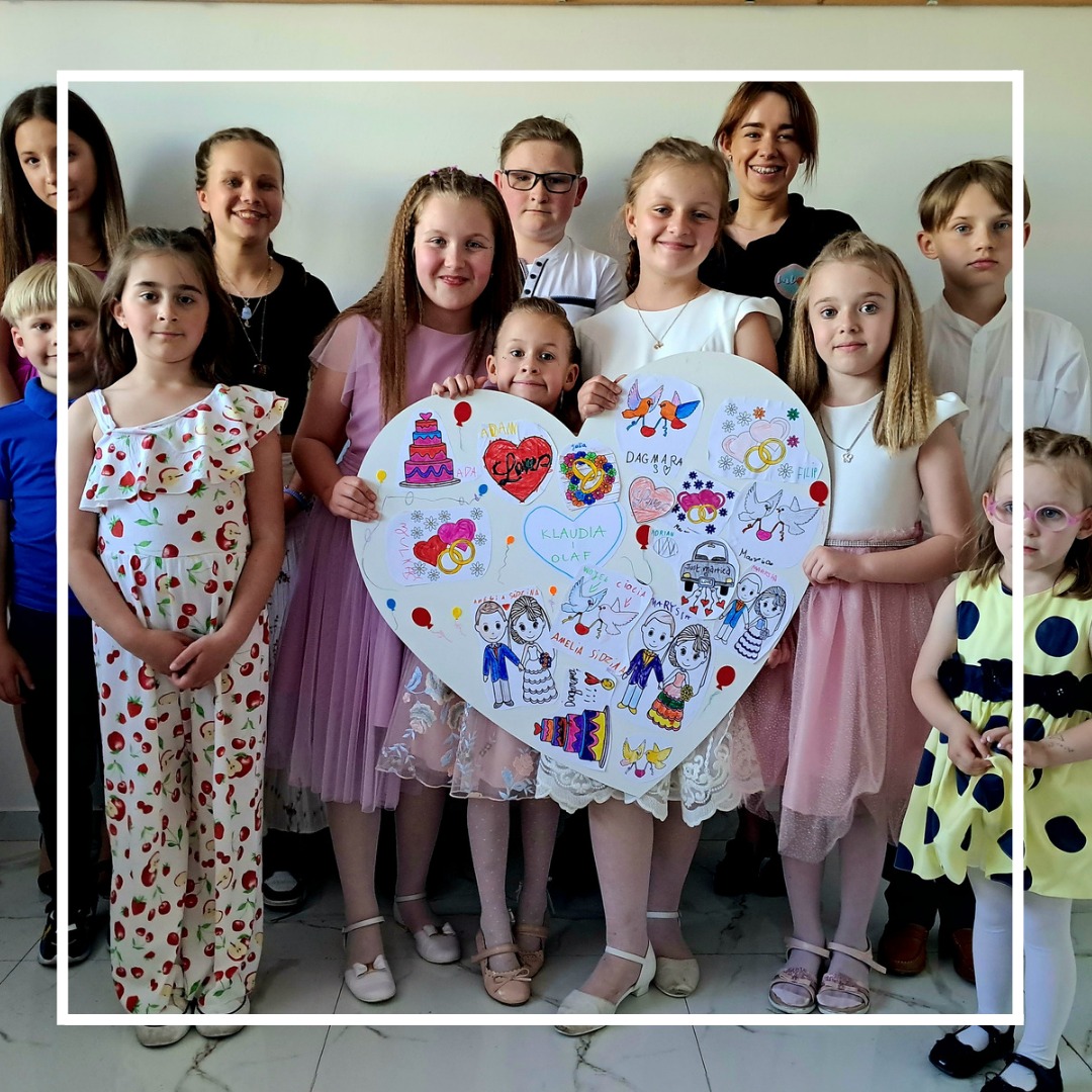 Grupa dzieci trzymająca duże, ręcznie wykonane serce z rysunkami i imionami, prawdopodobnie prezent dla pary młodej.