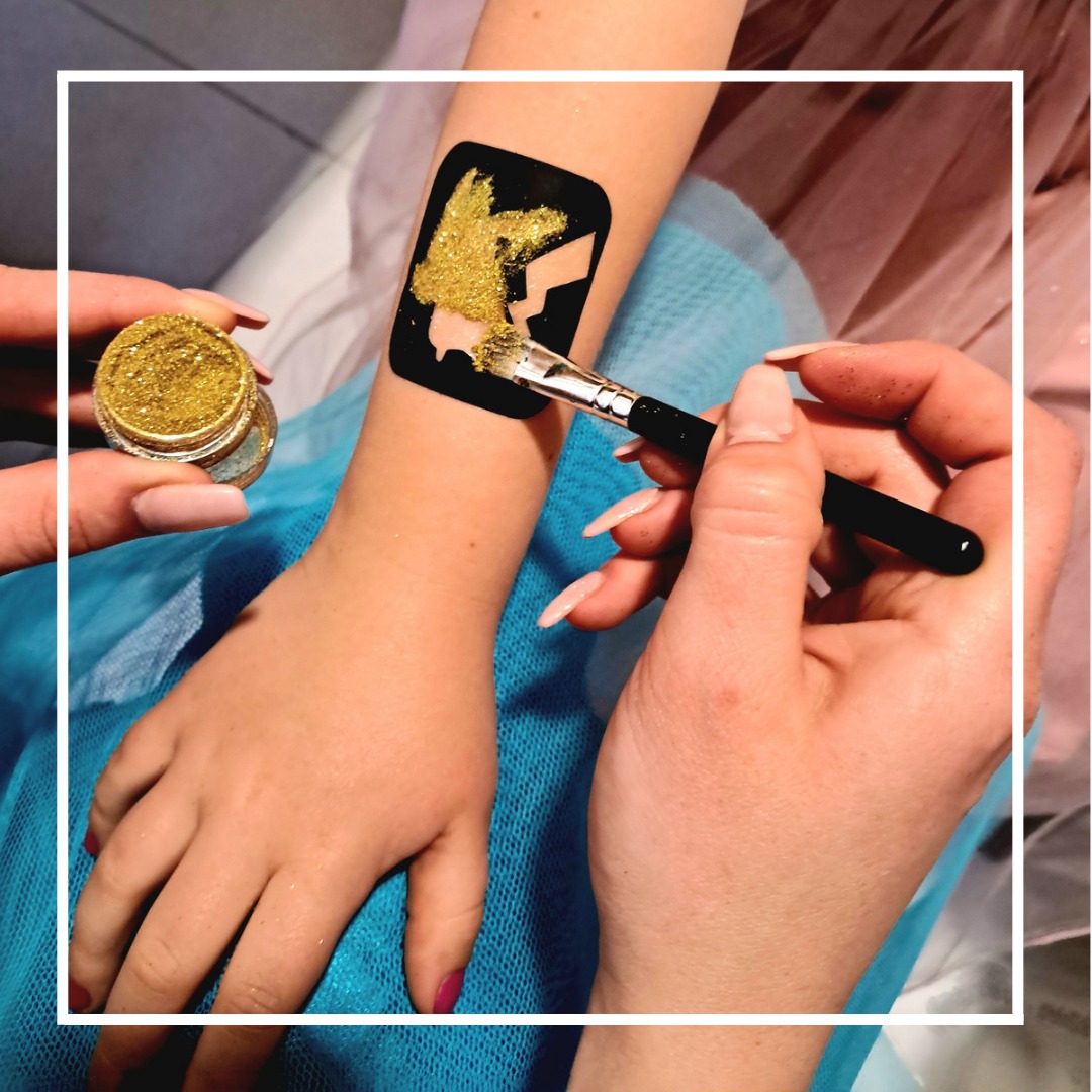 Nakładanie złotego brokatu na szablon Pikachu na ramieniu dziecka za pomocą pędzla; słoiczek brokatu w lewym górnym rogu.