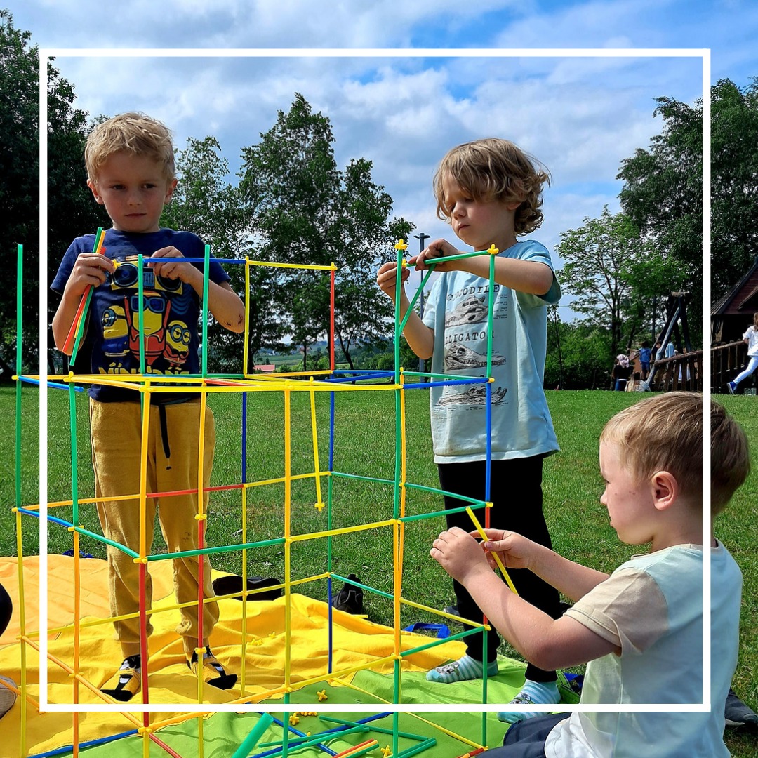 Trójka dzieci buduje konstrukcję z kolorowych rurek na żółtej macie na trawie w plenerze, w tle drzewa i niebieskie niebo z chmurami.