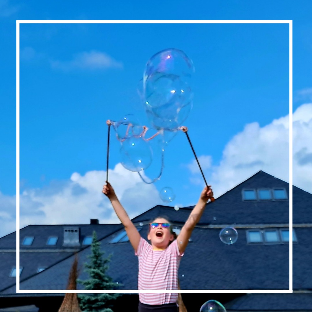 Dziewczynka w różowo-białej koszulce i okularach przeciwsłonecznych, z otwartymi ustami, trzyma nad głową przyrząd do puszczania ogromnych baniek mydlanych na tle błękitnego nieba i dachu domu.
