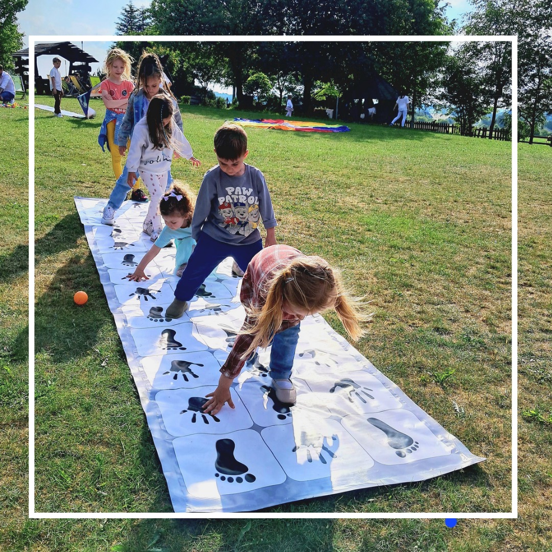 Grupa dzieci bawi się na trawie, grając w grę z odciskami dłoni i stóp na białej macie, z pomarańczową piłką obok, w tle widoczne osoby i kolorowa płachta.