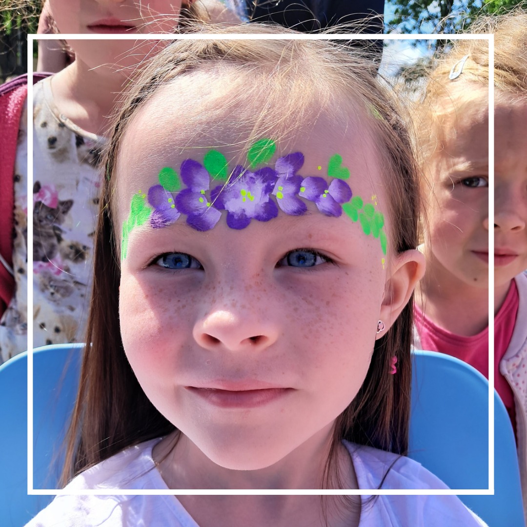 Dziewczynka z fioletowo-zielonymi kwiatami wymalowanymi na czole, usługa wykonana w Radomsku, w tle widoczne fragmenty twarzy innych dzieci.