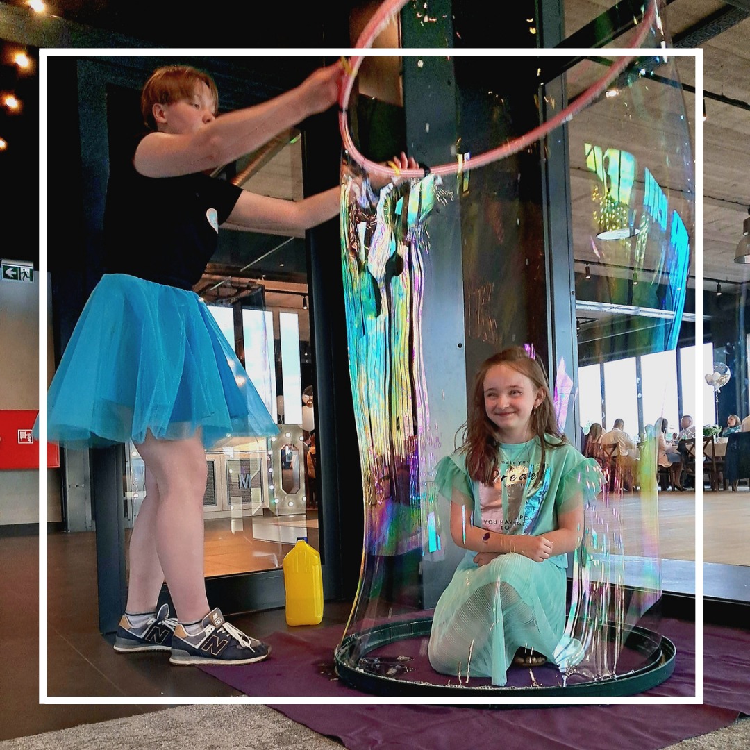 Animator w niebieskiej spódnicy tworzy ogromną bańkę mydlaną, w której siedzi uśmiechnięta dziewczynka; w tle widoczni ludzie przy stolikach.