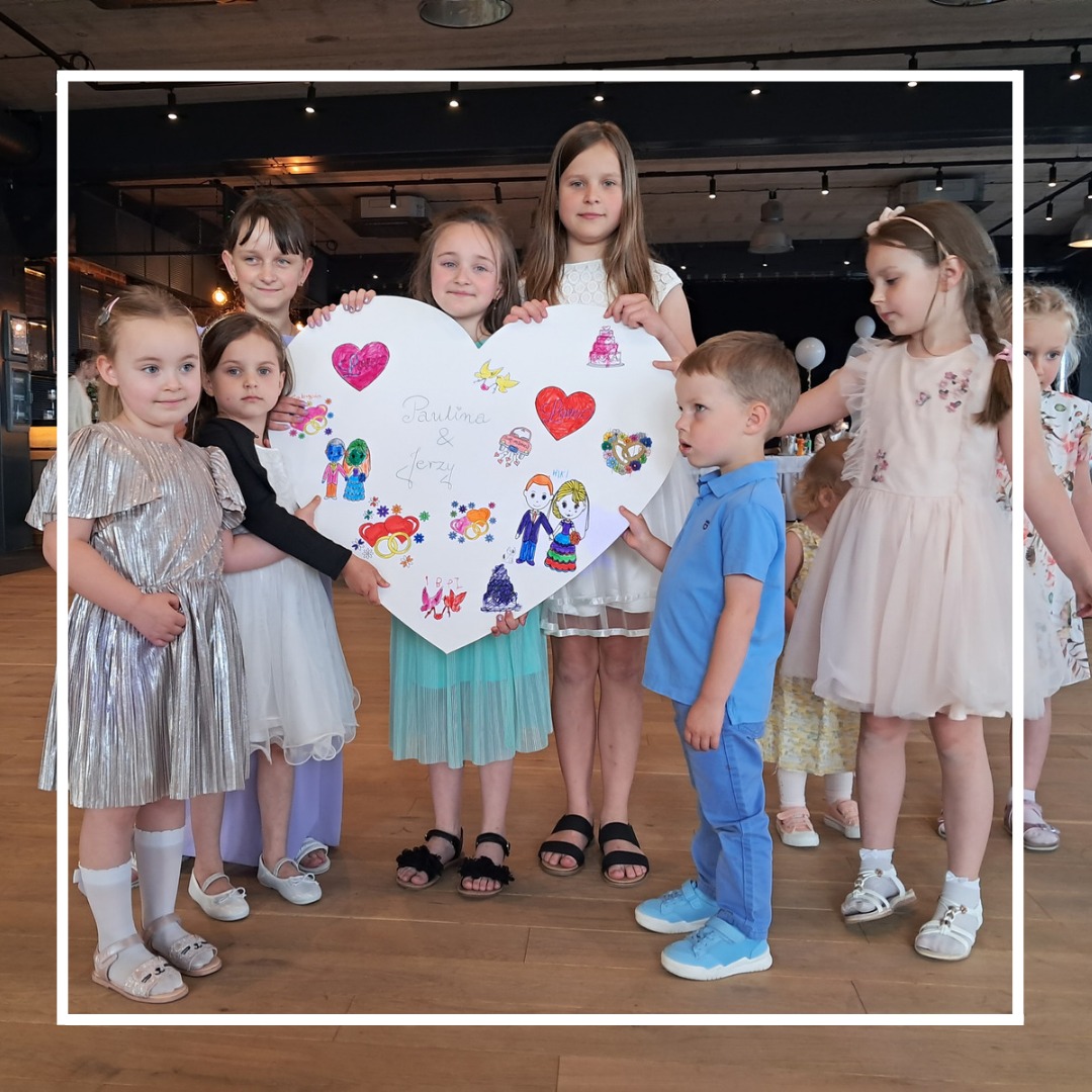 Grupa dzieci trzymająca duży karton w kształcie serca z rysunkami i napisem 'Paulina & Jerzy' podczas uroczystości, prawdopodobnie wesela.