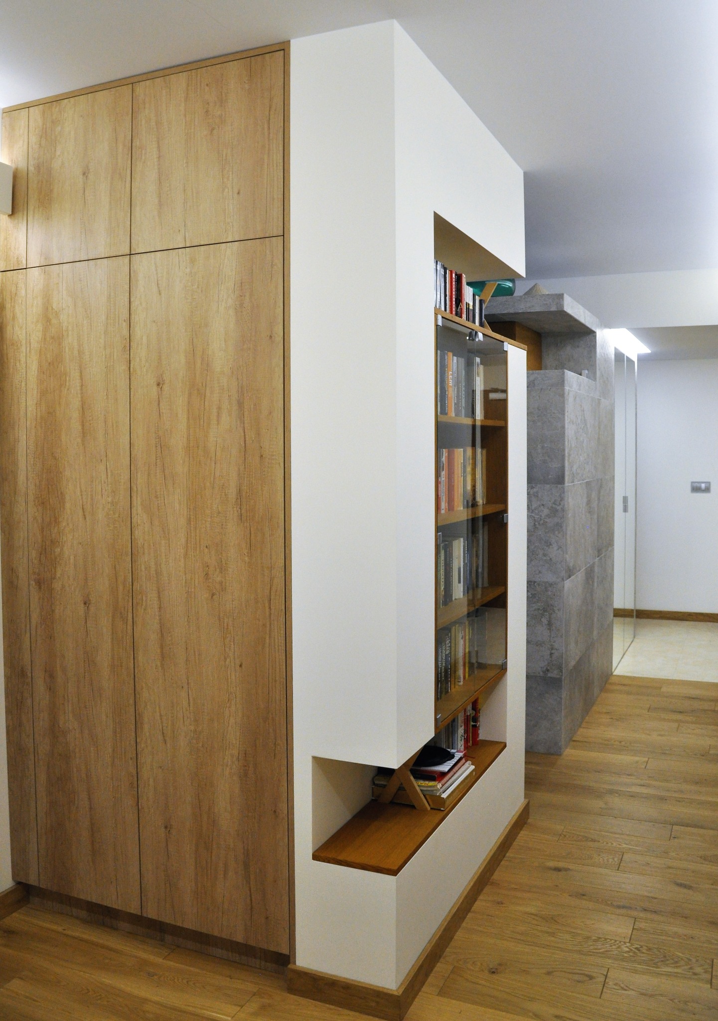 Wbudowana szafa z drewnianymi frontami sąsiadująca z białą zabudową regału z przeszkloną witryną na książki, widok na korytarz z szarą ścianą z płytek.