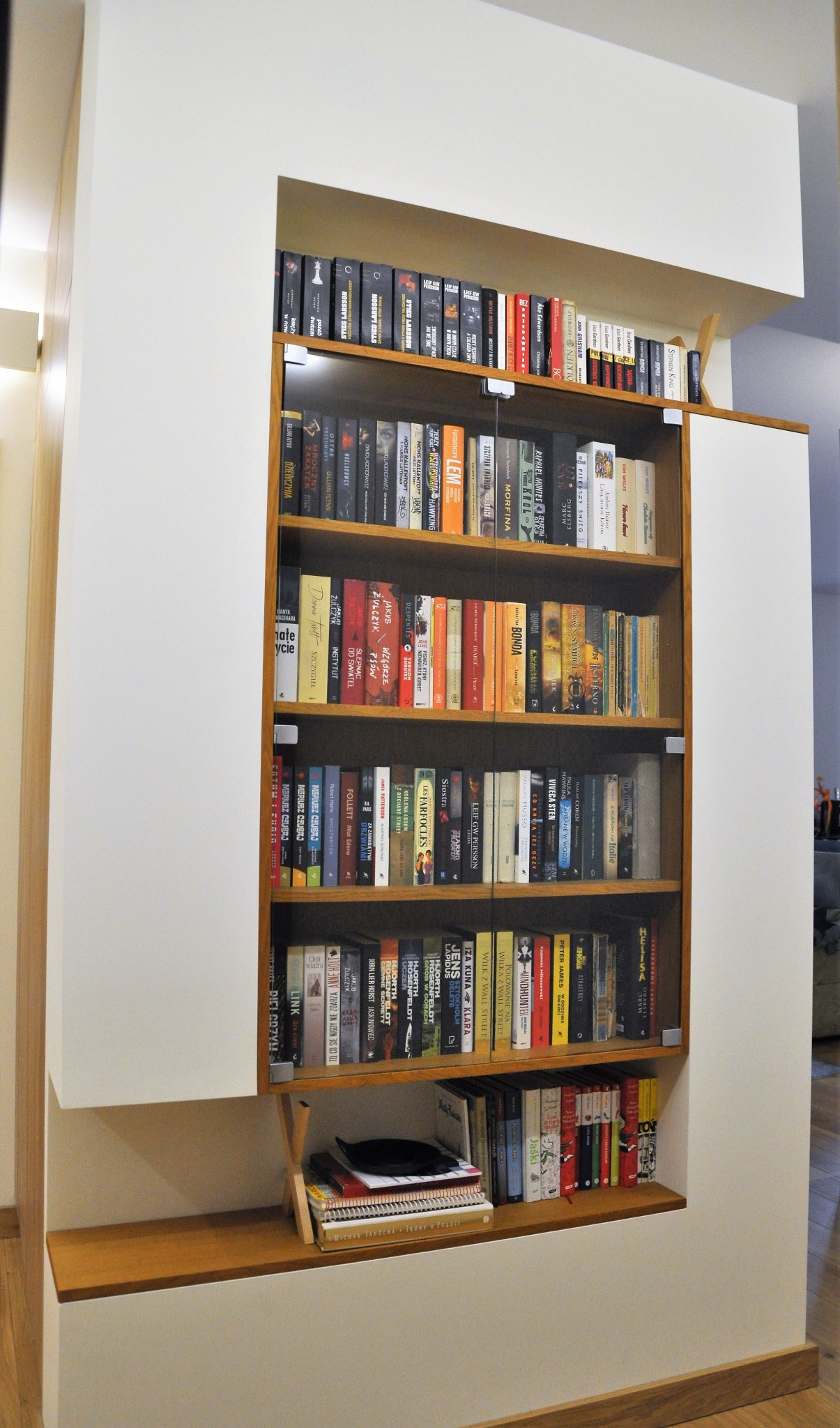 Zabudowana biblioteczka z półkami wypełnionymi książkami, notatnikami i czarną miseczką, wbudowana w białą ścianę z drewnianymi elementami wykończeniowymi.