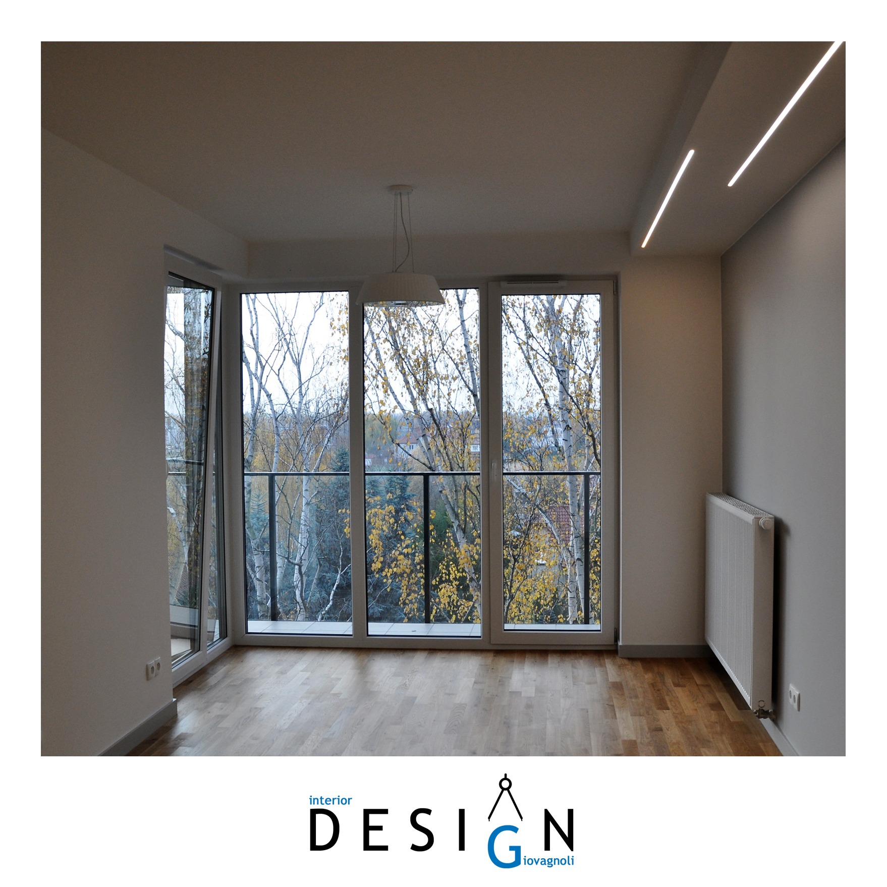 Jasne, puste wnętrze mieszkania z dużymi oknami balkonowymi wychodzącymi na drzewa z jesiennymi liśćmi, parkiet na podłodze, biały grzejnik i nowoczesne oświetlenie sufitowe.