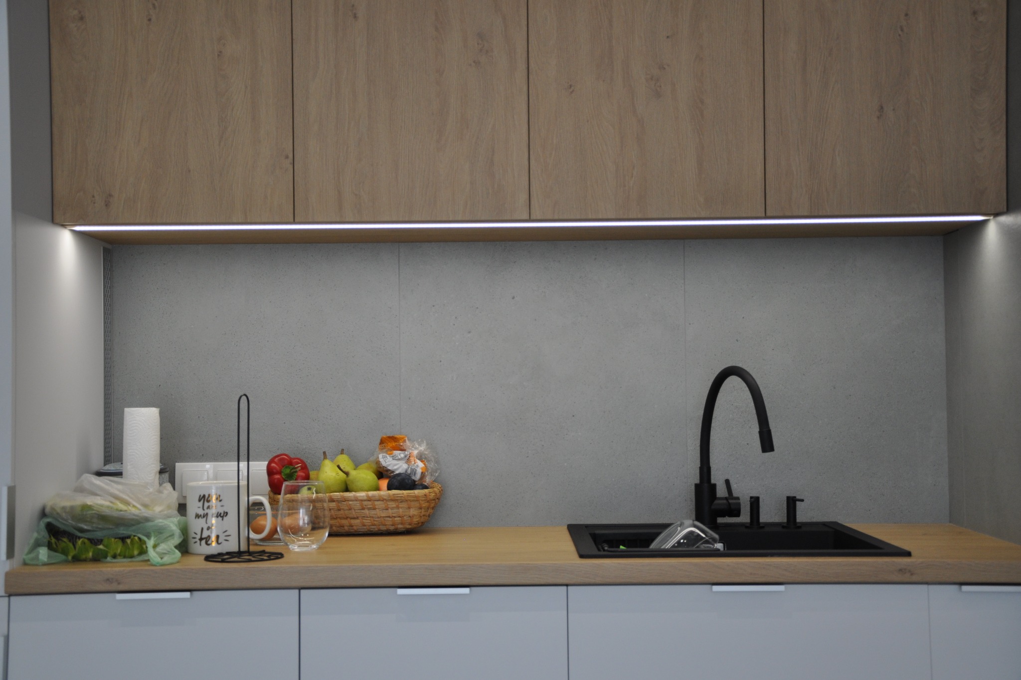 Nowoczesna kuchnia w Warszawie z drewnianymi szafkami górnymi podświetlonymi listwą LED, szarym panelem ściennym i blatem z czarnym zlewem oraz koszem owoców. Na blacie widoczny także kubek...
