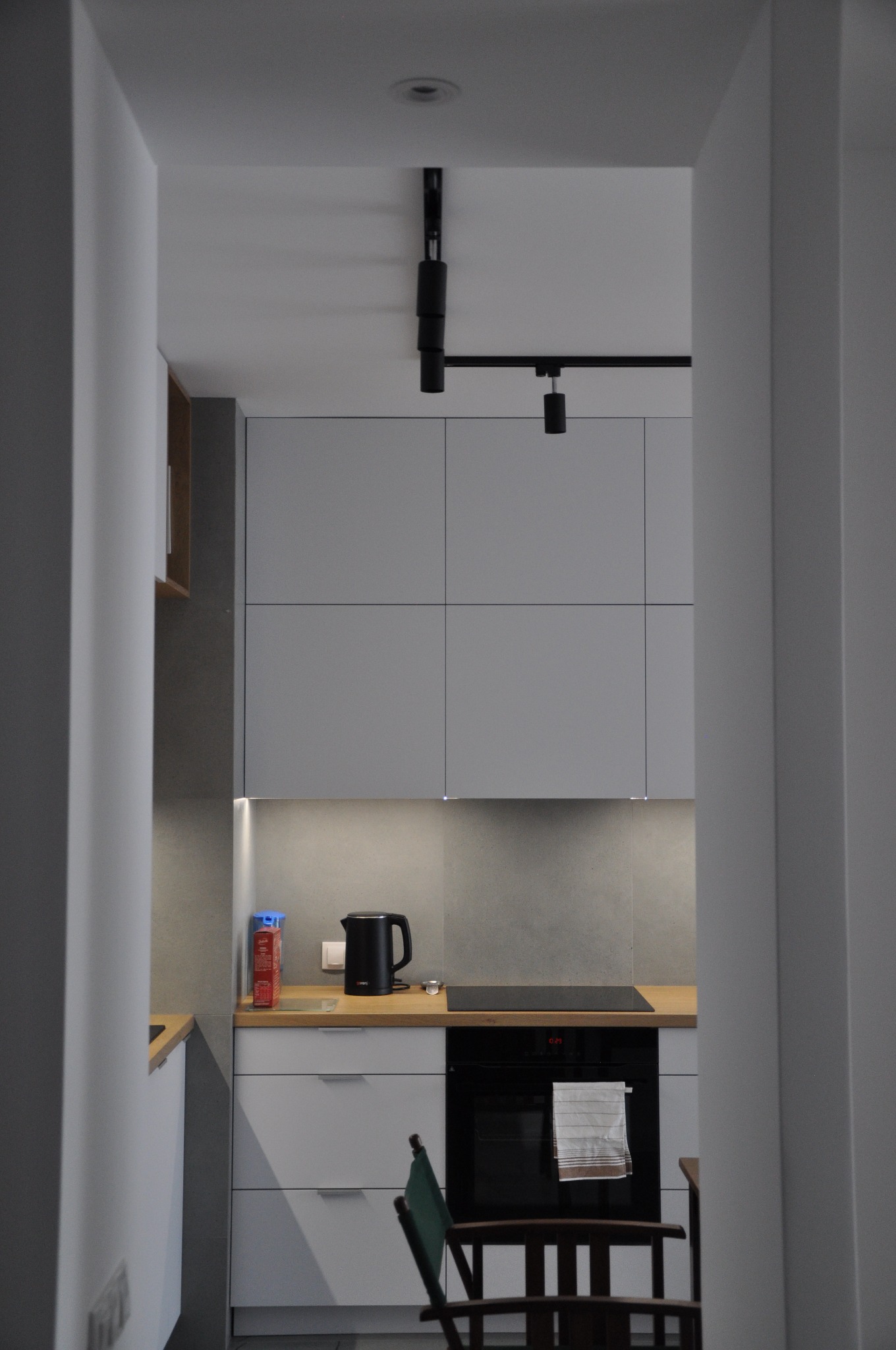 Minimalistyczna kuchnia w odcieniach szarości z drewnianym blatem, oświetleniem podszafkowym LED i czarnymi lampami sufitowymi na szynie, widziana zza framugi drzwi.