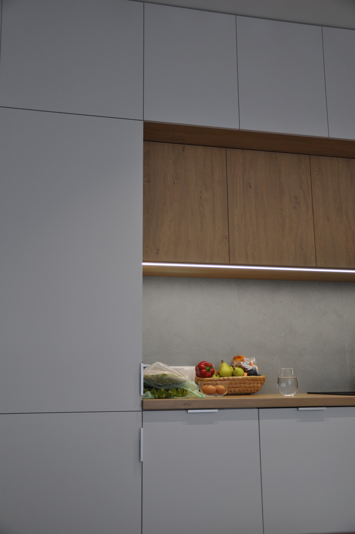 Nowoczesna kuchnia z szarymi frontami szafek i drewnianymi akcentami, podświetlona listwą LED, na blacie koszyk z owocami, warzywami i jajkami oraz szklanka z wodą.