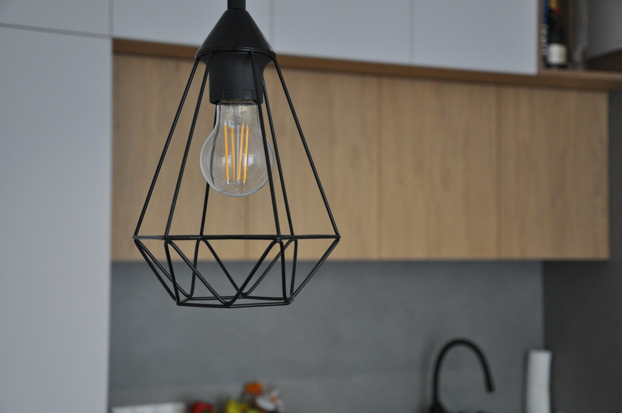 Czarna, geometryczna lampa wisząca z żarówką filamentową na tle drewnianych szafek kuchennych i szarej ściany.