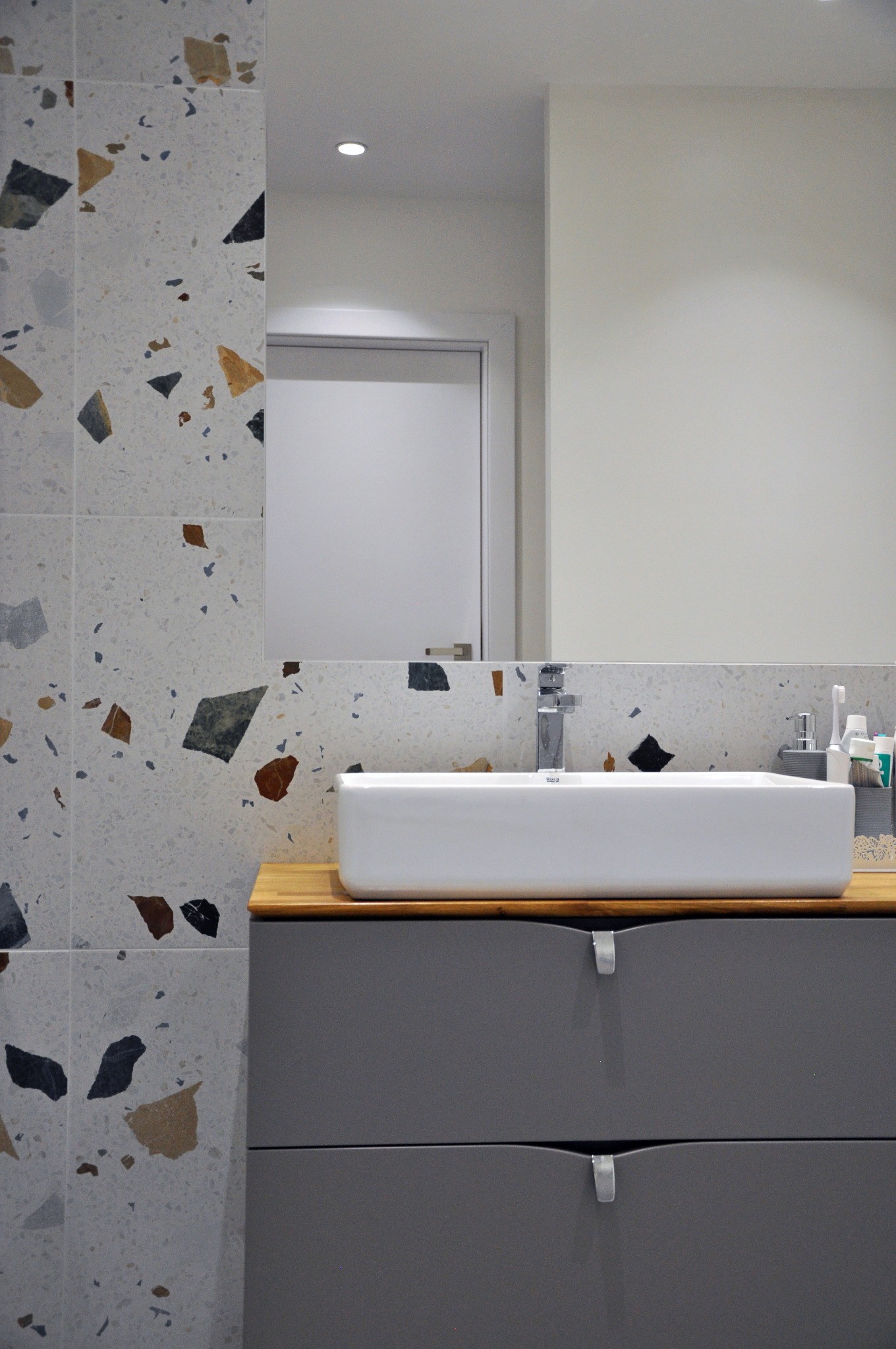 Nowoczesna łazienka z umywalką nablatową, szarą szafką z dwoma szufladami i płytkami imitującymi lastryko na ścianie, odbicie drzwi w lustrze.