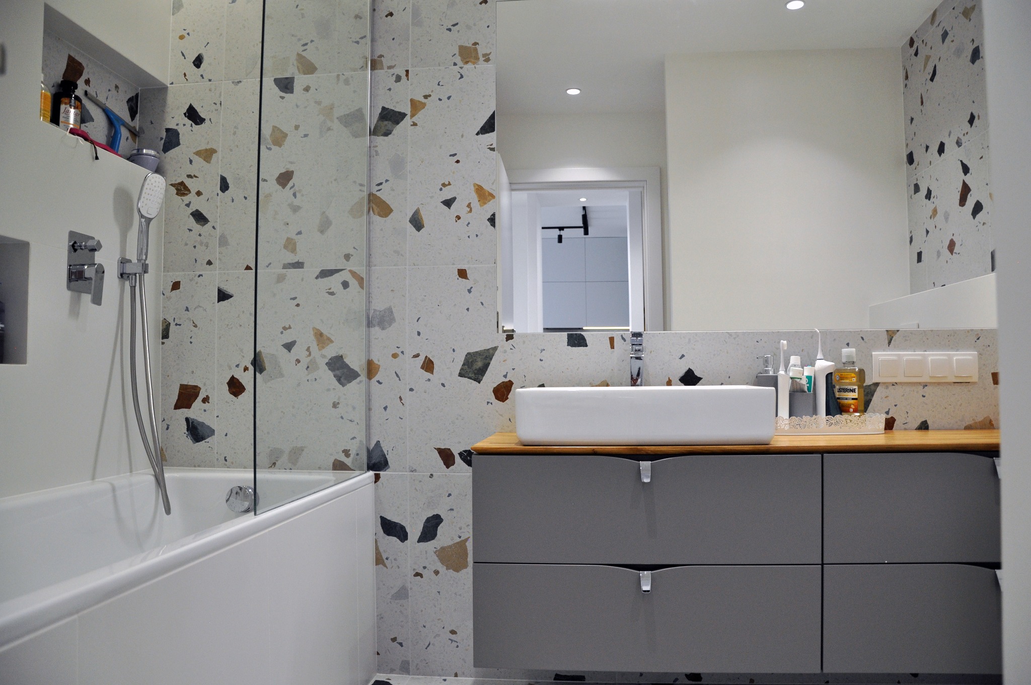 Nowoczesna łazienka z wanną i szklanym parawanem, umywalką nablatową na szafce z szufladami w odcieniach szarości, płytki imitujące lastryko na ścianach.