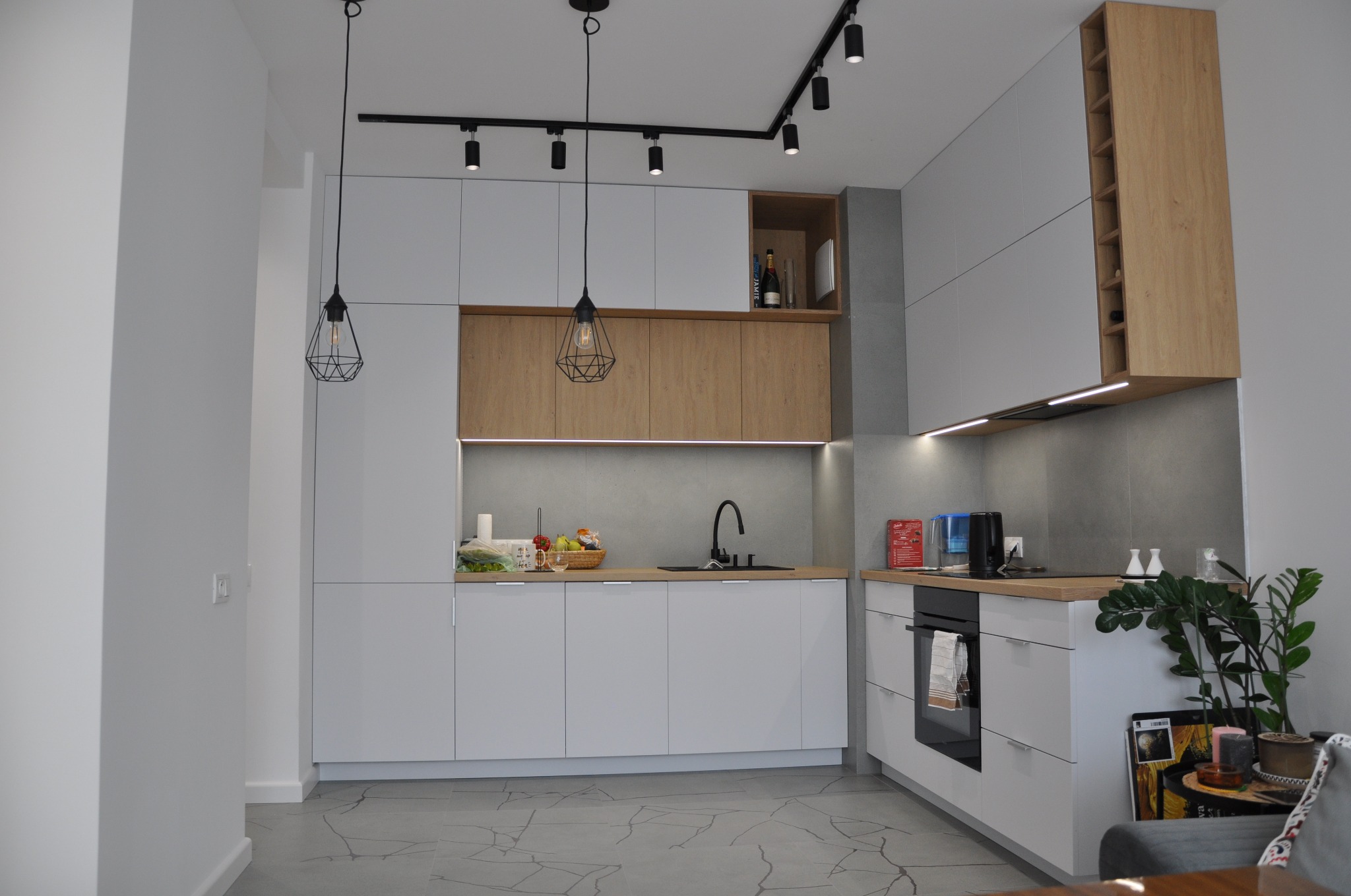 Nowoczesna kuchnia w odcieniach szarości i drewna, z wiszącymi lampami o geometrycznych kloszach i punktowym oświetleniem szynowym, uzupełniona o drewniane akcenty w zabudowie i blatach.
