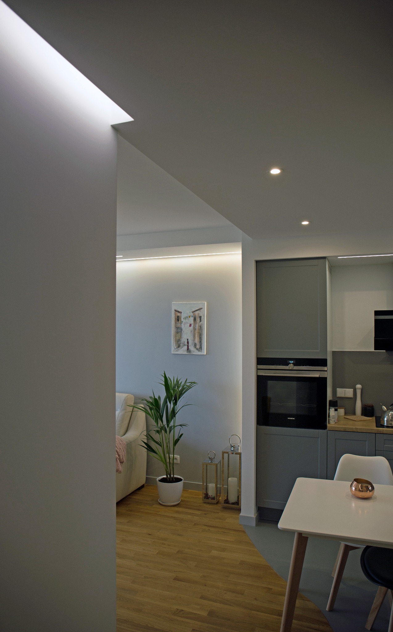Nowoczesne wnętrze mieszkania z szarą zabudową kuchenną, drewnianą podłogą, rośliną doniczkową i subtelnym oświetleniem LED w suficie.