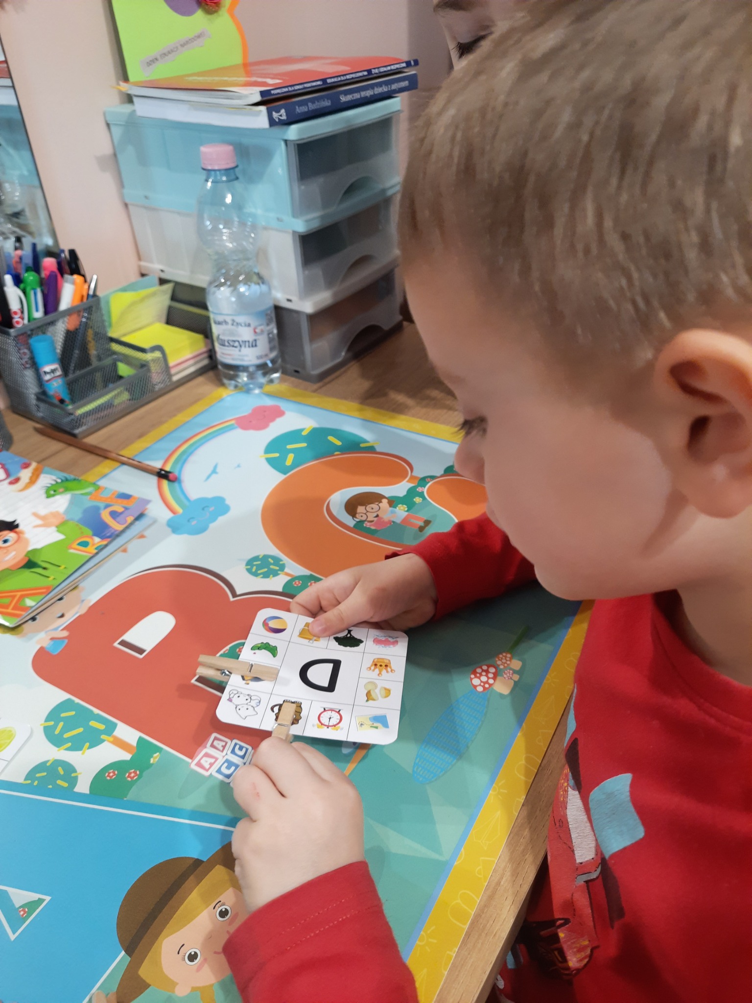 Chłopiec w czerwonej bluzce ćwiczy wymowę litery D z pomocą kart obrazkowych i klamerek na biurku z kolorową planszą edukacyjną.