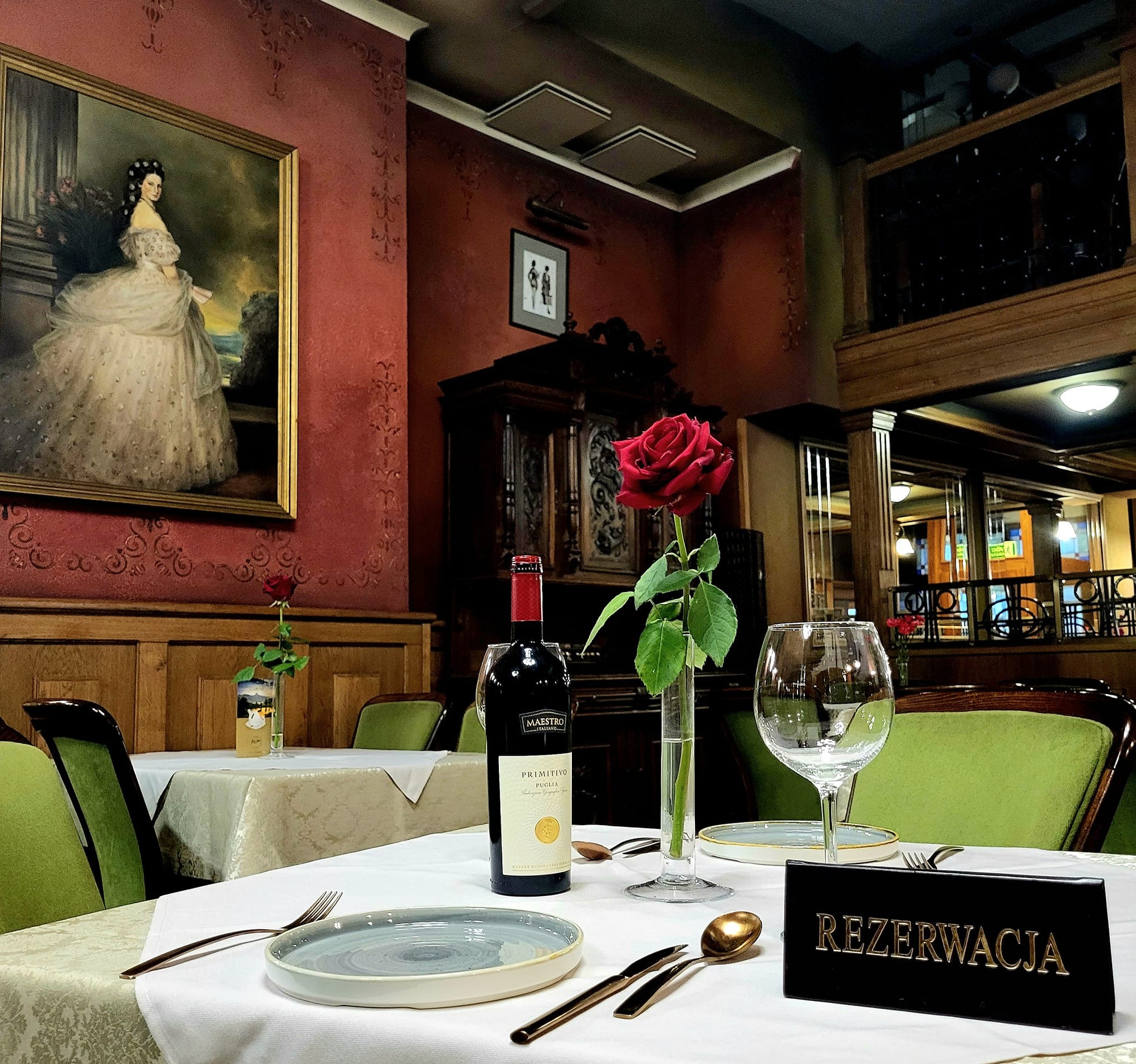 Uroczysty stół w restauracji z rezerwacją, butelką wina Maestro Primitivo, pojedynczą czerwoną różą w wazonie, zastawą stołową i obrazem damy w balowej sukni na ścianie w tle.