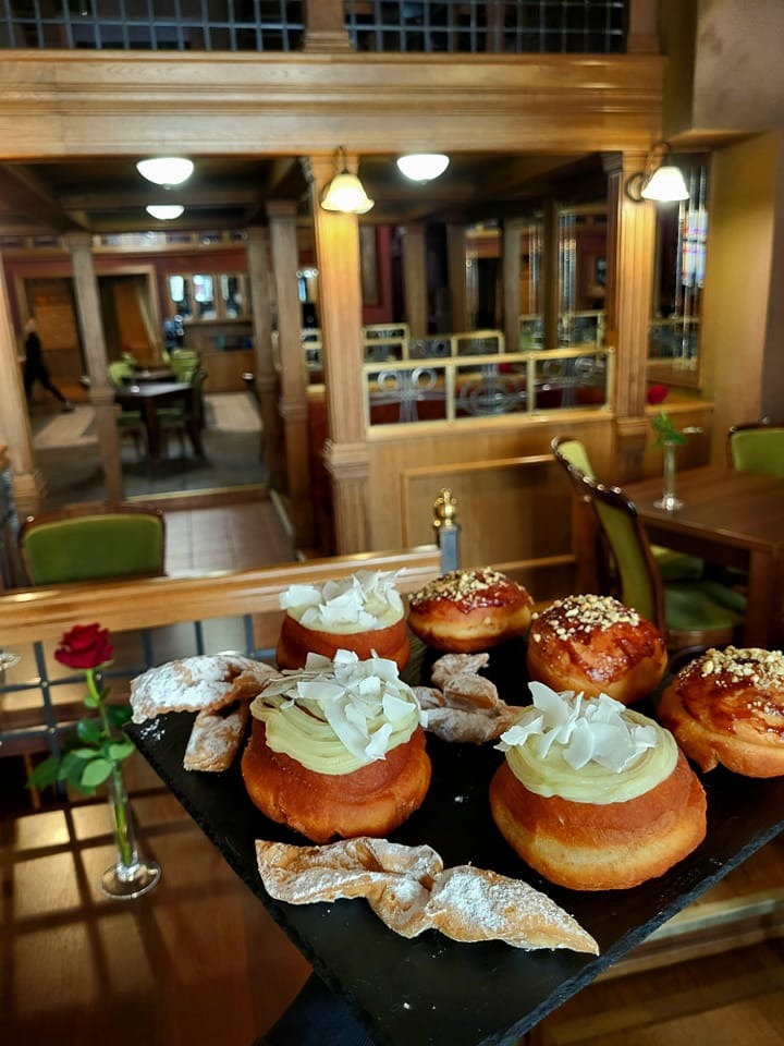 Taca z pączkami z kremem, posypką i faworkami oprószone cukrem pudrem, na tle eleganckiego wnętrza restauracji z drewnianą balustradą i stolikami.