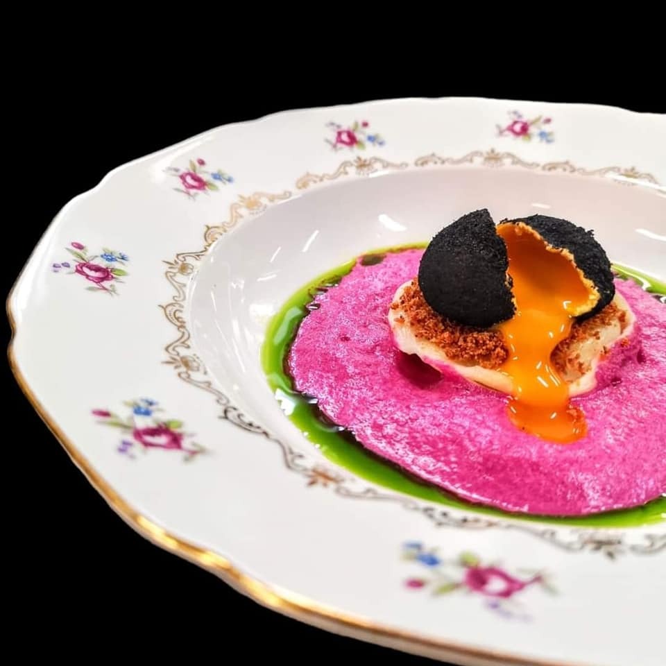 Elegancko podane danie: różowe puree z zielonym sosem, na nim biały element posypany brązowym proszkiem i czarna kula z wypływającym pomarańczowym sosem, całość na talerzu w stylu vintage...