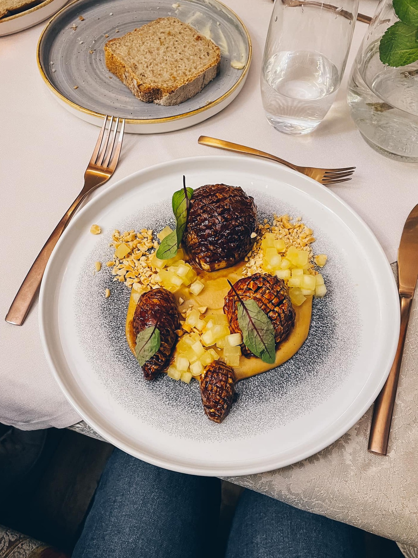 Elegancko podane danie z trzema mini szyszkami, puree, kostkami ananasa i posiekanymi orzechami na białym talerzu z szarą obwódką, obok złote sztućce, chleb i woda w szklance.