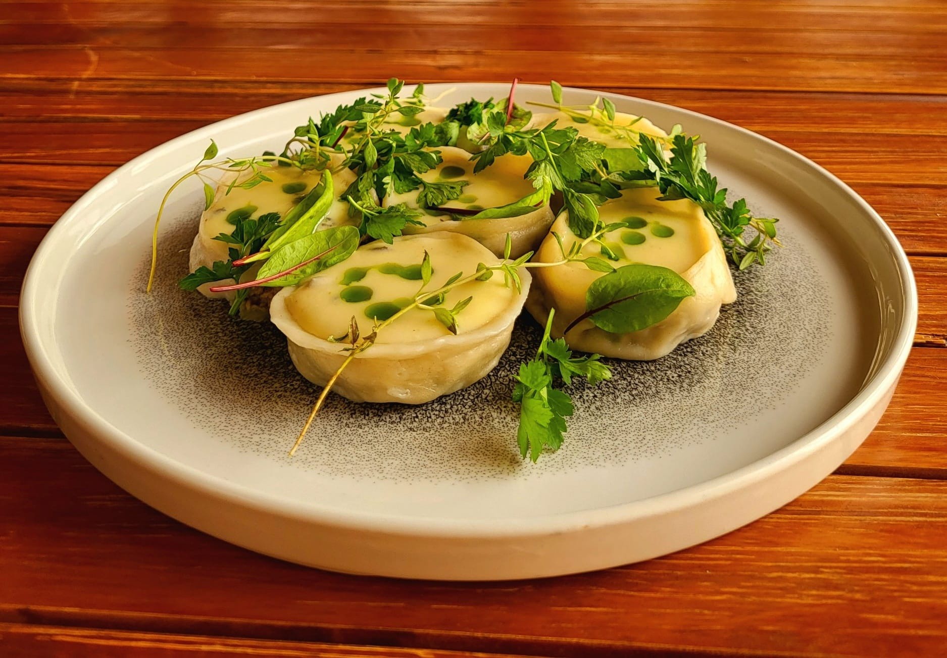Elegancko podane pierożki z farszem, polane sosem i udekorowane świeżymi ziołami na talerzu z delikatnym wzorem, całość umieszczona na drewnianym stole.