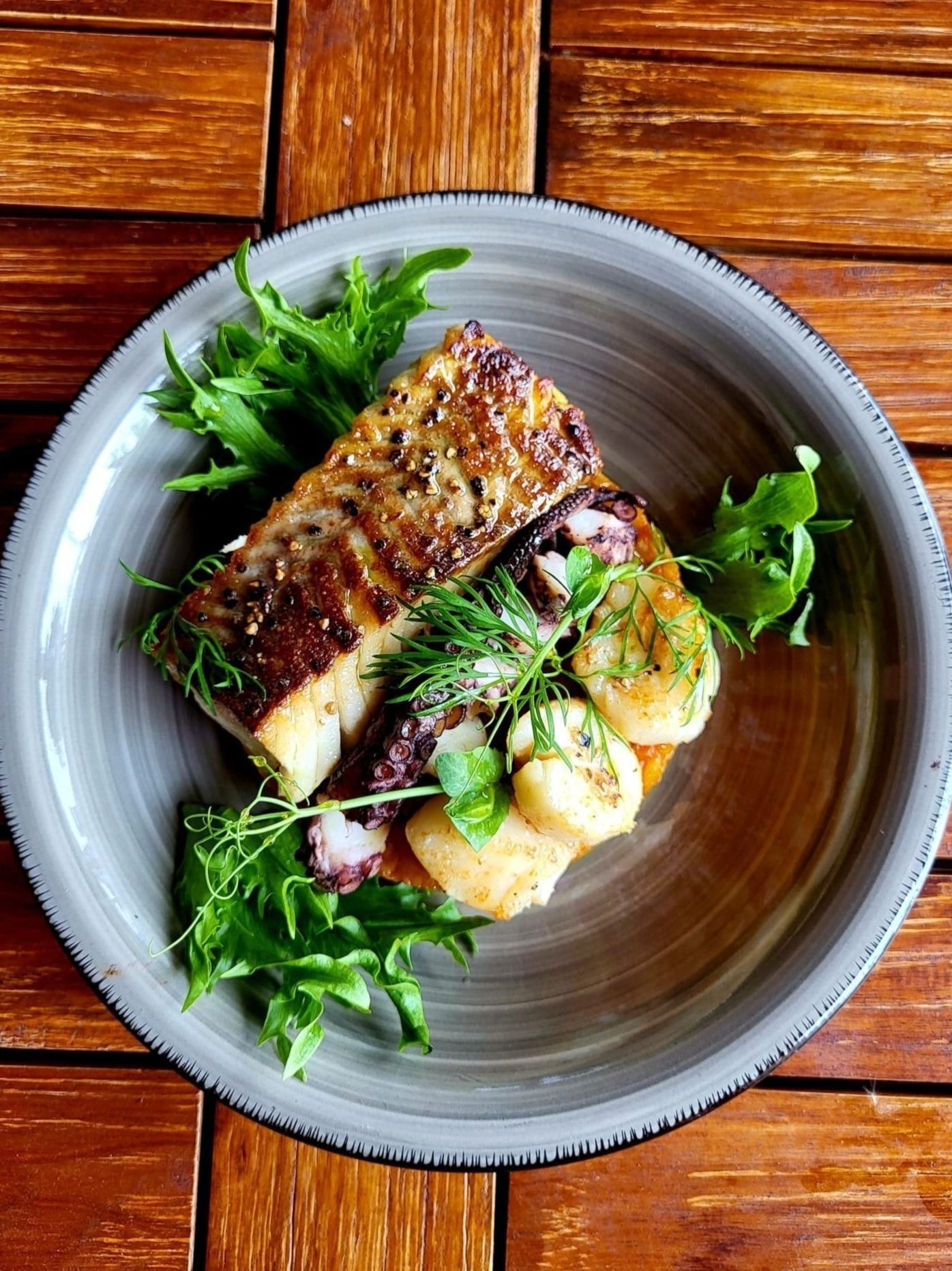 Filet z ryby z grilla, ośmiornica i przegrzebki na talerzu z rukolą i koperkiem, serwowane na drewnianym stole.