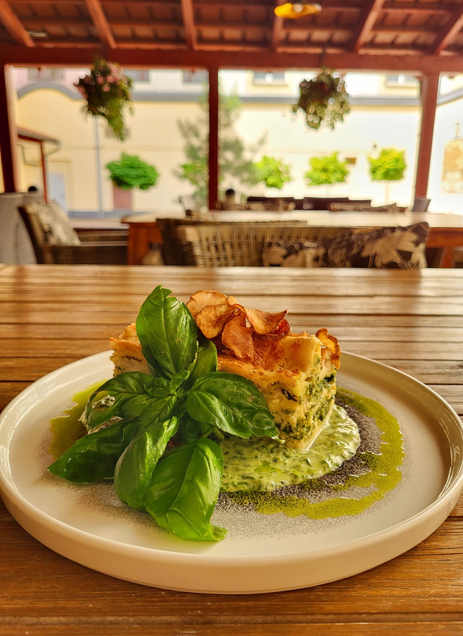 Elegancko podana porcja zielonej lasagne z bazylią i chipsami na talerzu, na drewnianym stole w restauracji z widokiem na podwórze.