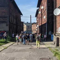 Grupa turystów idzie ulicą między ceglanymi budynkami w postindustrialnej dzielnicy, widoczne grafitti na ścianach, satelity na budynkach.