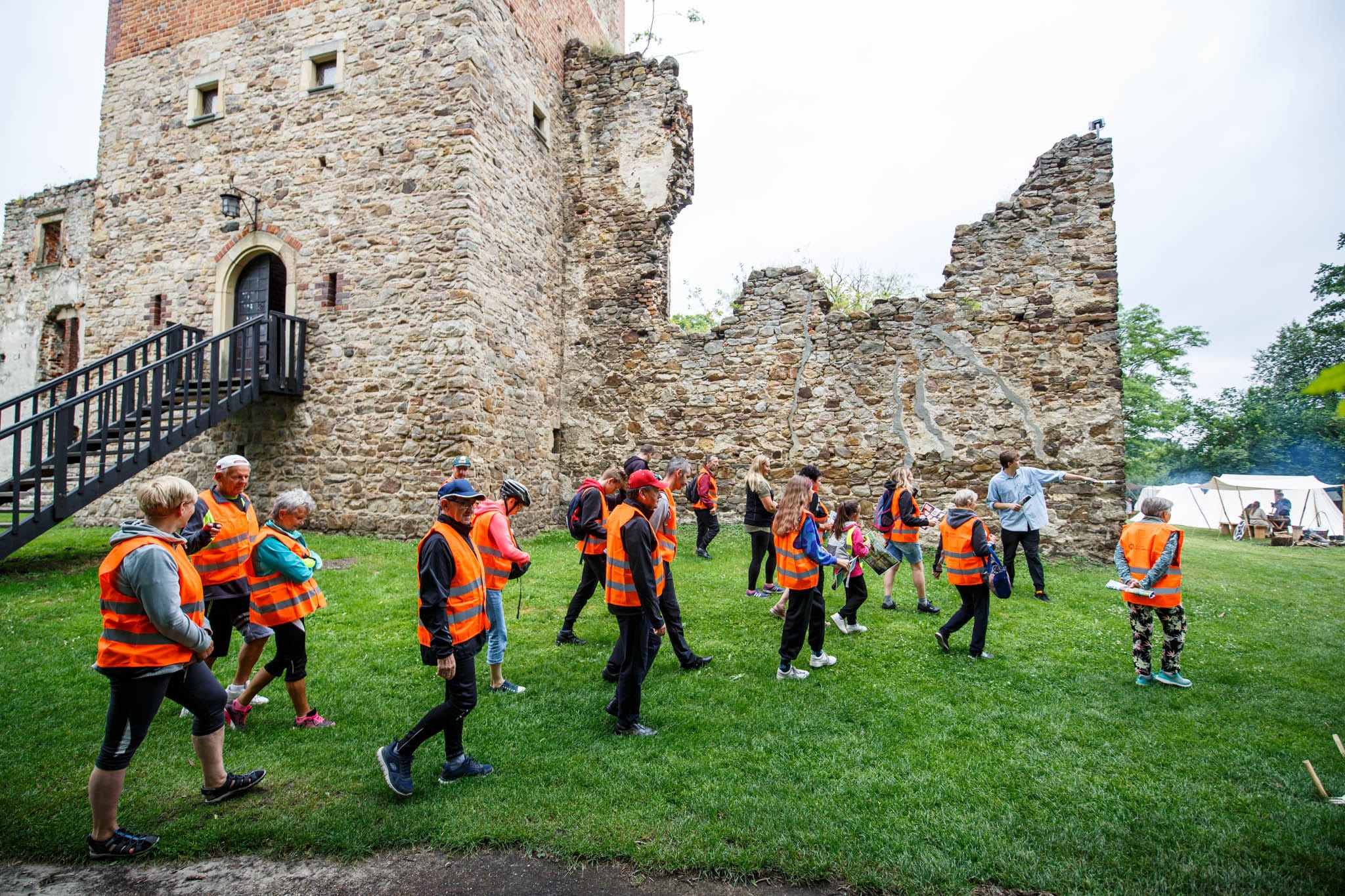 Grupa turystów w pomarańczowych kamizelkach zwiedza ruiny średniowiecznego zamku; przewodnik wskazuje na fragment muru, w tle rozstawione namioty obozowiska.