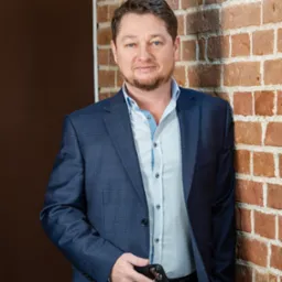 Portret adwokata w niebieskim garniturze, opierającego się o ceglaną ścianę, trzymającego telefon w dłoni.