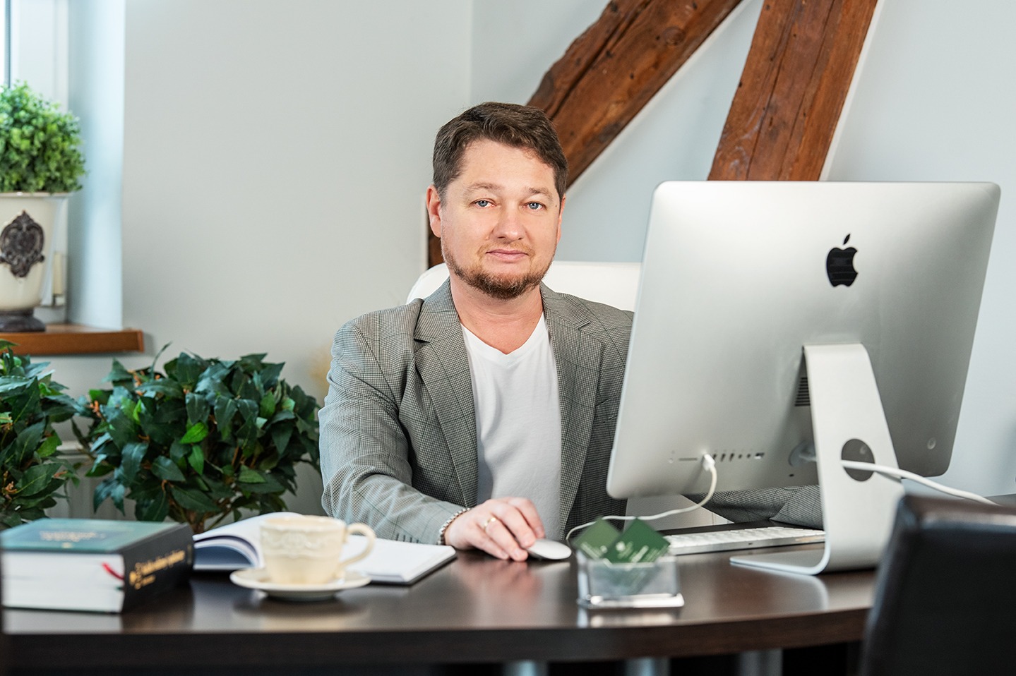Portret adwokata w biurze, siedzącego przy biurku z komputerem iMac, filiżanką kawy, notatnikiem i książkami, z widoczną drewnianą belką stropową w tle.