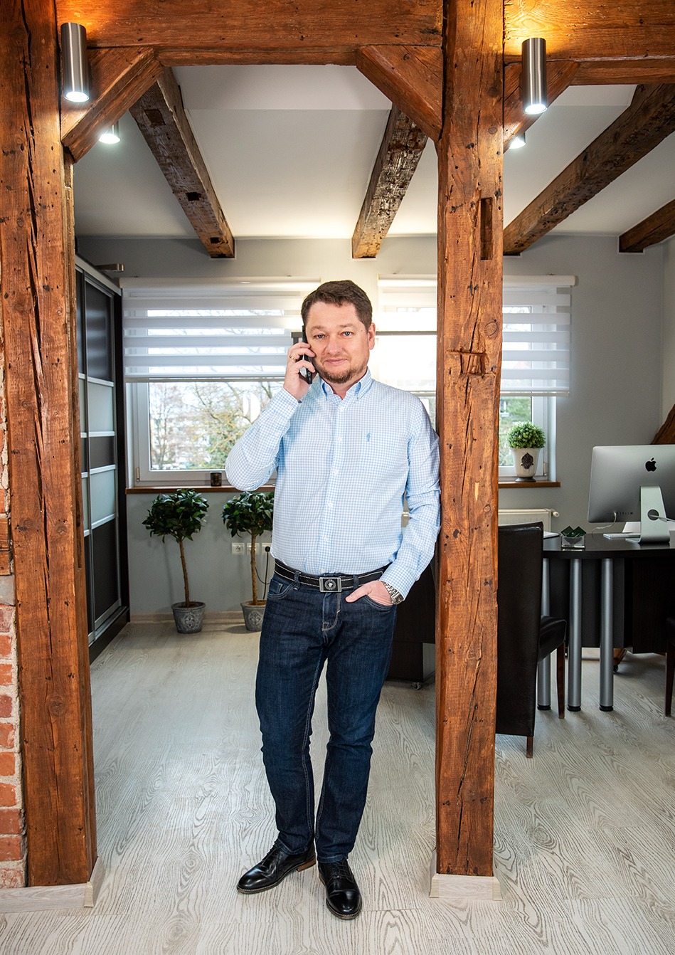 Mężczyzna w niebieskiej koszuli rozmawia przez telefon, stojąc w biurze z drewnianymi belkami i ceglanym elementem, w tle biurko z komputerem.