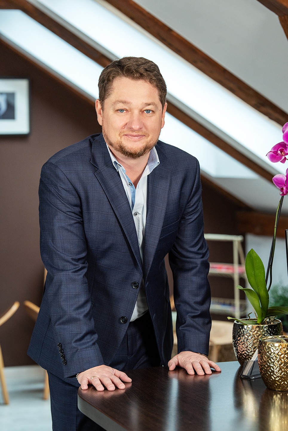 Uśmiechnięty mężczyzna w granatowym garniturze, oparty o ciemny stół z dekoracyjnymi doniczkami i storczykiem, w tle skośny sufit z drewnianymi belkami.