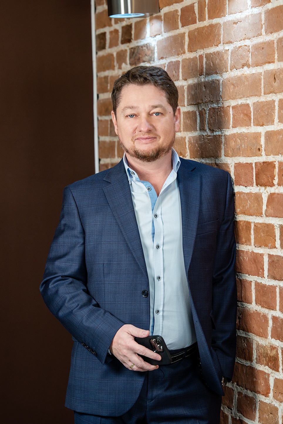 Portret adwokata w niebieskim garniturze, opierającego się o ceglaną ścianę, trzymającego telefon w dłoni.