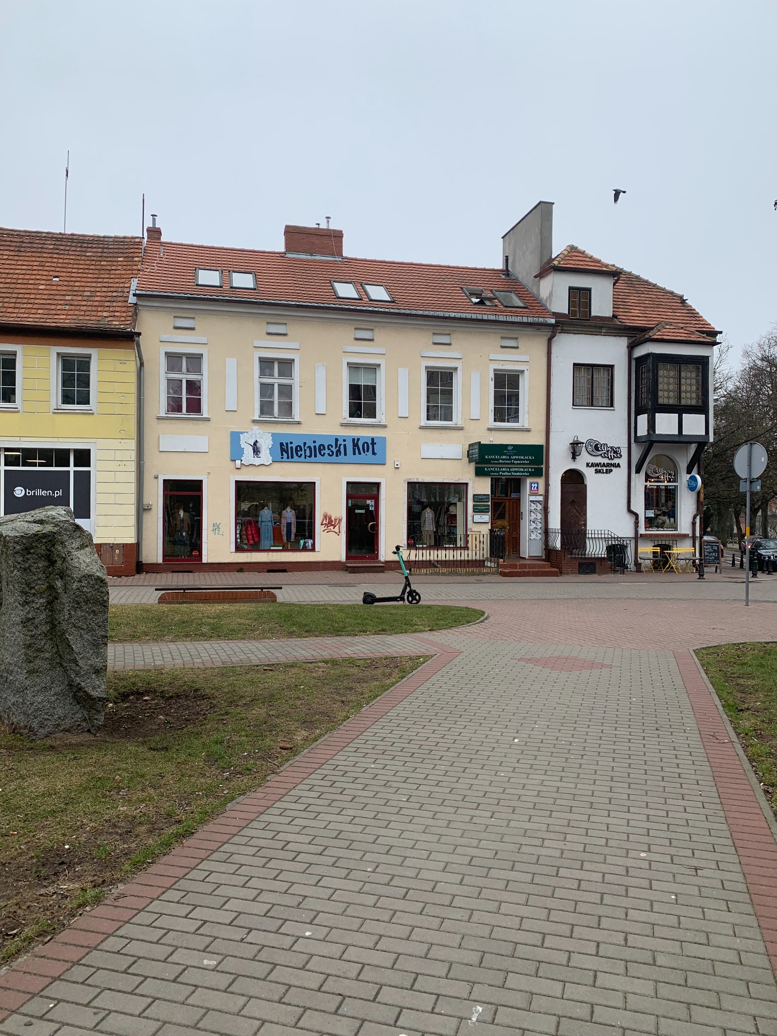 Ulica z kolorowymi kamienicami, w tym budynek z szyldem 'Kancelaria Adwokacka' i 'Kawiarnia Sklep', widok z chodnika.