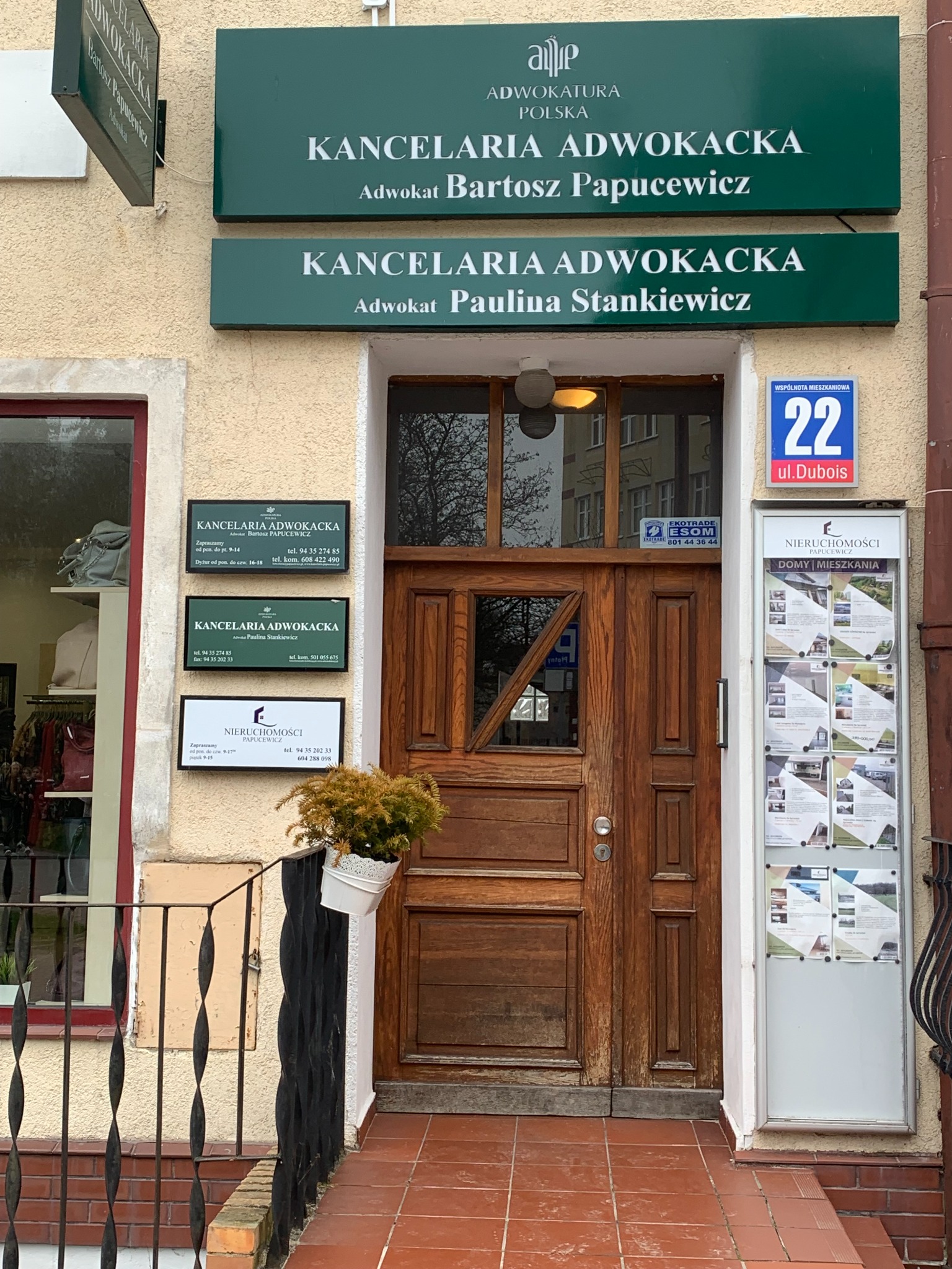 Wejście do kancelarii adwokackiej z drewnianymi drzwiami i szyldami informacyjnymi na fasadzie budynku, donica z rośliną przy barierce.