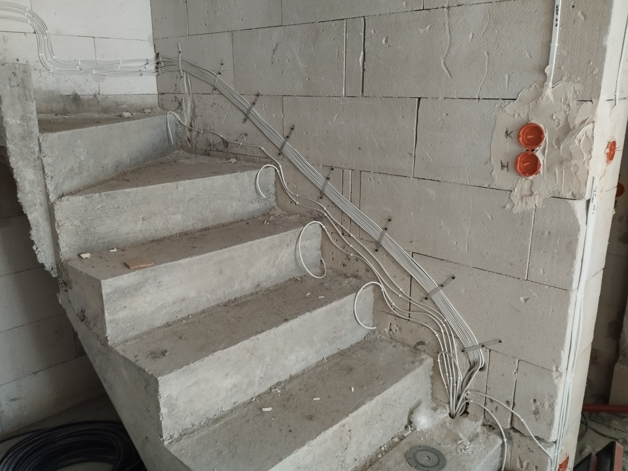 Surowe schody z instalacją elektryczną w peszlach, zamocowane do ściany z bloczków betonu komórkowego, widoczne puszki instalacyjne.