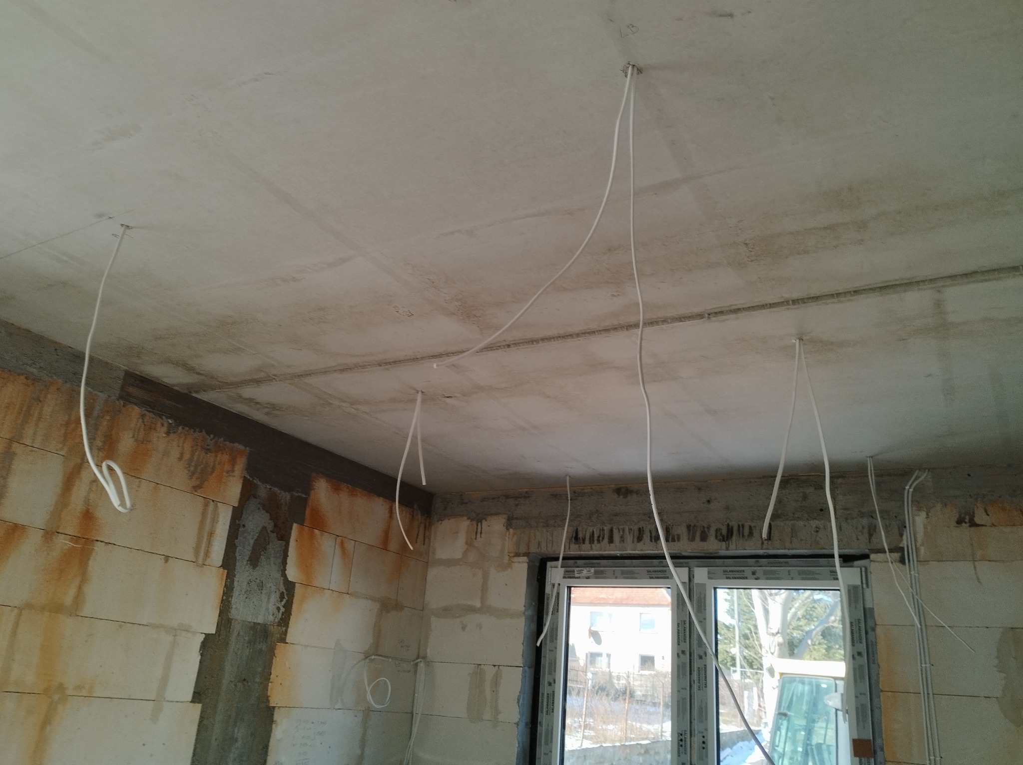 Surowy strop z wyprowadzonymi przewodami elektrycznymi w nowo budowanym pomieszczeniu z oknem. Widoczne bloczki betonowe.