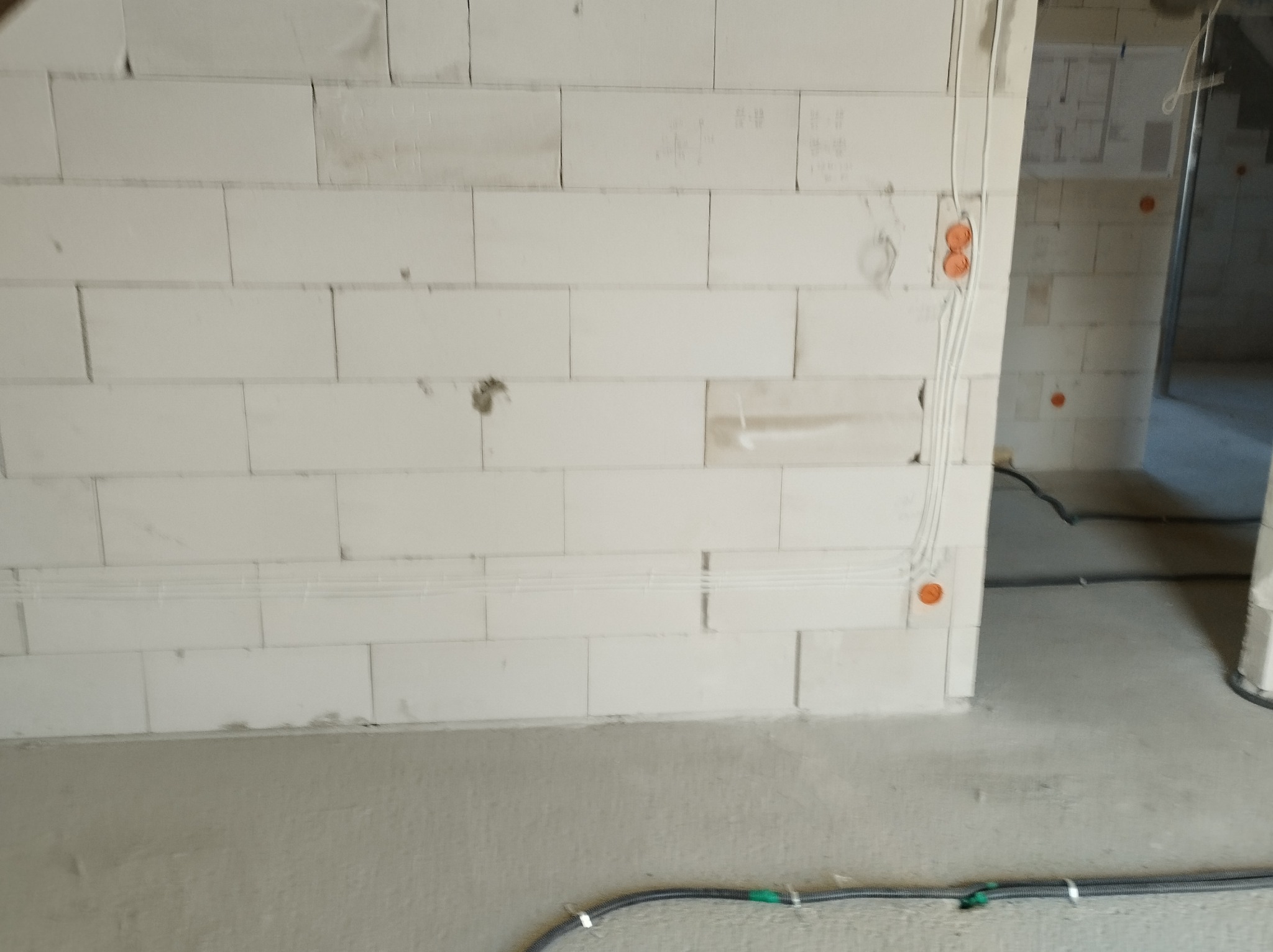 Ściana z betonu komórkowego z widocznymi przewodami elektrycznymi, pomarańczowe puszki instalacyjne. W tle widoczny plan budynku na ścianie.