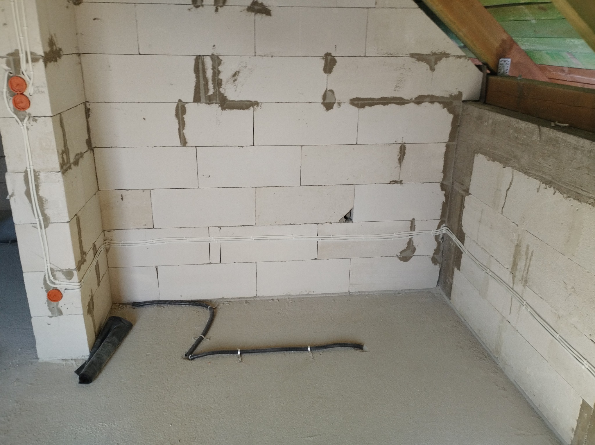 Surowy stan poddasza w Knybawie: białe bloczki, instalacja elektryczna w rurkach, puszki i peszel na podłodze. Widoczne elementy konstrukcji dachu.