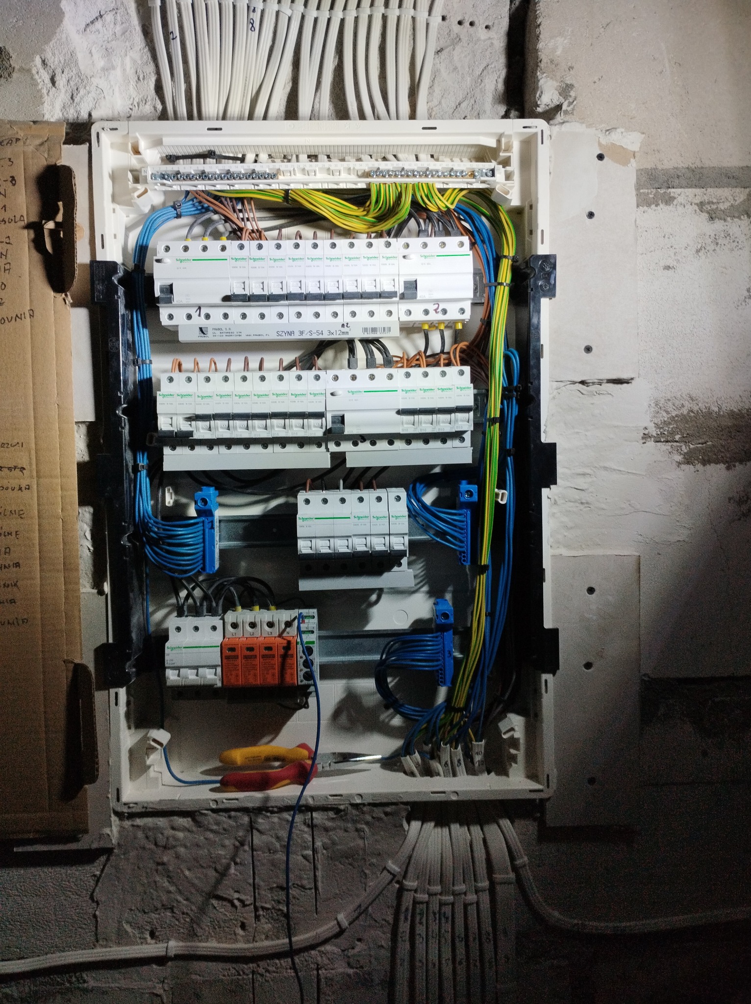 Rozdzielnia elektryczna Schneider z precyzyjnie ułożonymi przewodami w kolorach: niebieskim, żółto-zielonym i brązowym, zamontowana na surowej ścianie. Widoczne narzędzia.